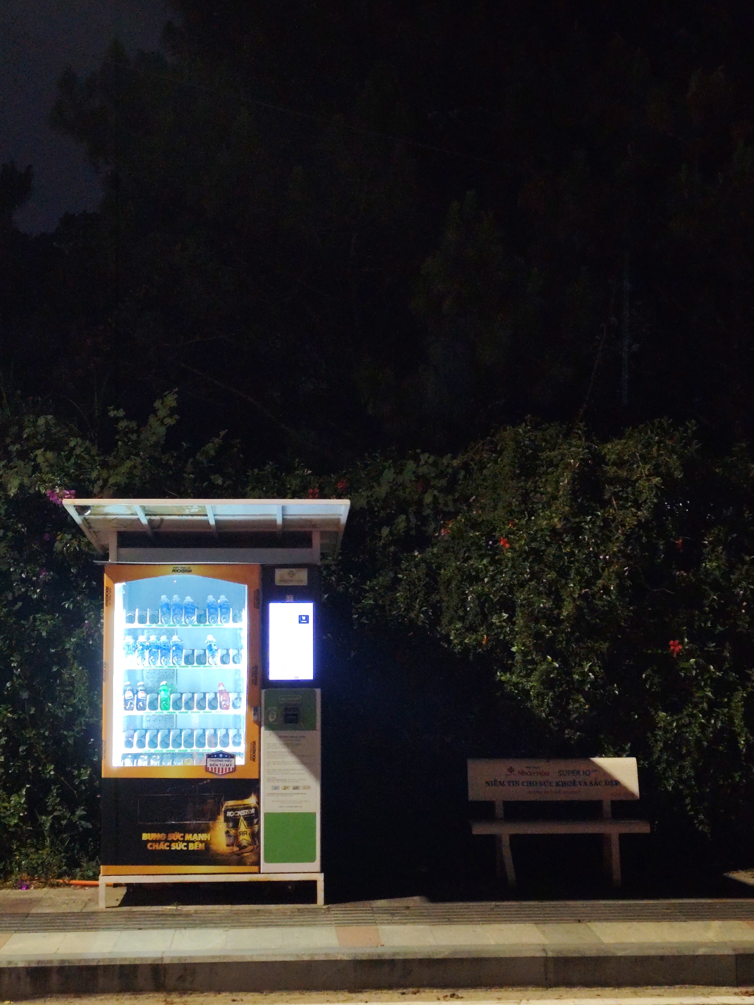 The image captures a nighttime scene in what appears to be Da Lat, Vietnam. A brightly lit vending machine stands prominently on the left, stocked with bottled drinks and canned beverages. The machine is branded with "ROCKSTAR" and Vietnamese text. To the right of the vending machine is a bench with Vietnamese text on the back, suggesting it's a public seating area. The background is dominated by dense, dark shrubbery with some visible red flowers, adding to the nighttime ambiance. The overall impression is a quiet, urban scene with a functional vending machine contrasting against the natural, overgrown environment. The location is indicated by the text on the bench mentioning "NHTHOU SUPER IQ" and "NIỀM TIN CHO SỨC KHỎI VÀ SẮC ĐẸP", and the brand on the vending machine.