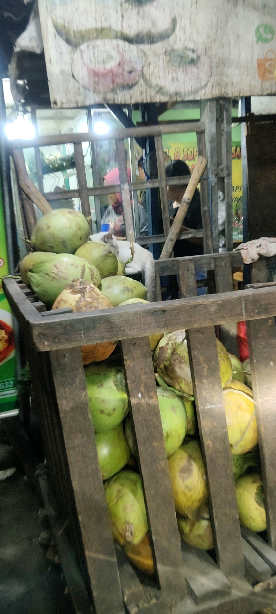 A cart full of green coconuts is displayed at a street vendor stall in Mekarmukti, Indonesia. The cart is made of weathered wood, with vertical slats on the sides. Several coconuts are stacked in the cart, with some at the top and others deeper within. The coconuts vary in size and color, with some being a vibrant green and others having brown or yellowish tinges. In the background, behind a metal grate, there are glimpses of people and products inside the stall. A white cat with its tail visible is partially obscured by coconuts in the cart. The scene appears to be outdoors, under a covered area with a sign visible above the cart.