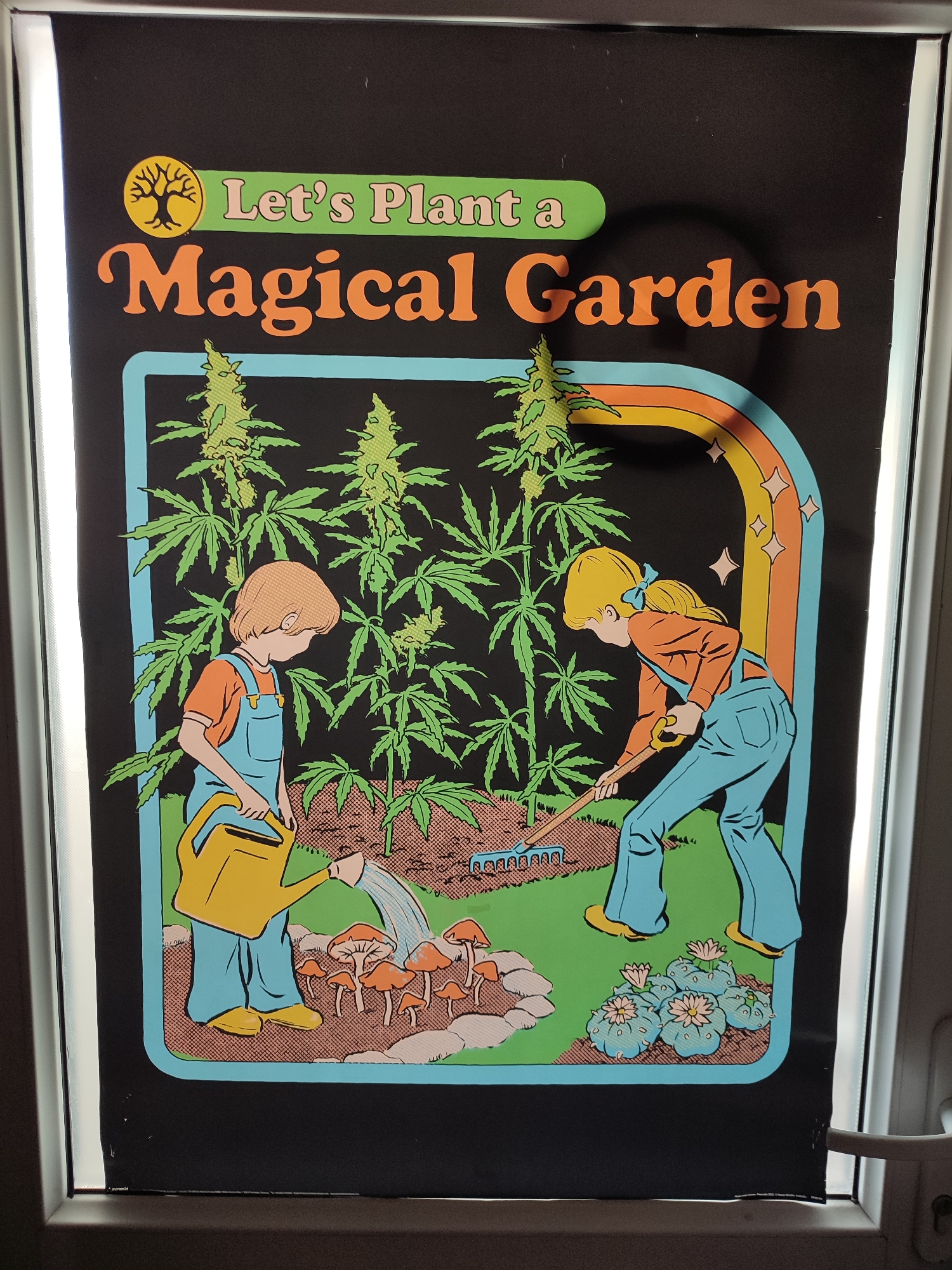The image shows a framed poster displayed on a window ledge. The poster features a stylized illustration with a black background. The title "Let's Plant a Magical Garden" is printed in a retro font at the top, accompanied by a yellow circular emblem with a tree silhouette. The central scene depicts two children tending to a garden of cannabis plants, mushrooms, and cactus-like plants. A boy in blue overalls waters the plants with a yellow watering can, while a girl in similar attire uses a rake. A rainbow with sparkling stars arches over the right side of the garden. The overall aesthetic is psychedelic and whimsical. The poster is framed in silver and appears to be lit from the front, creating some glare and shadows. The window frame and a white handle are visible on the right side of the image, indicating an indoor setting with a view of the outdoors, likely a residential or commercial space. There is no visible information within the image that places it in Üsküdar, Turkey.