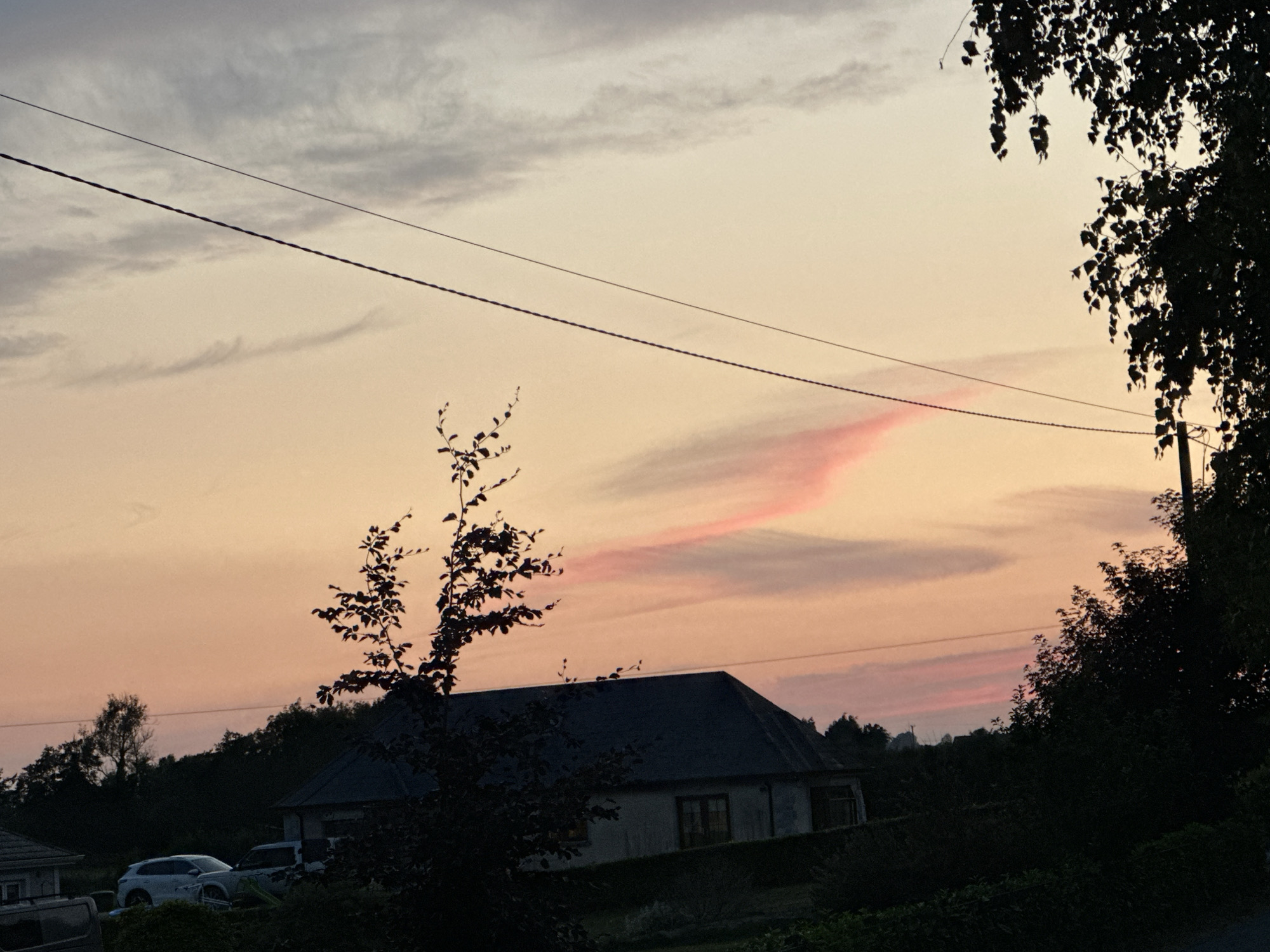 This image depicts a rural sunset scene in Moortown, Ireland, with a low-lying house and silhouetted trees dominating the foreground. The sky is a gradient of soft pinks and oranges, with wispy clouds that have caught the fading sunlight. Two power lines are visible, stretched horizontally across the sky, adding a man-made element to the natural landscape. In the lower portion of the image, the dark silhouette of a house with a pitched roof is visible, partially obscured by dense trees. Two cars, a white SUV and another vehicle, are parked in front of the house. The overall impression is one of quietude and the tranquil beauty of the Irish countryside at dusk.
