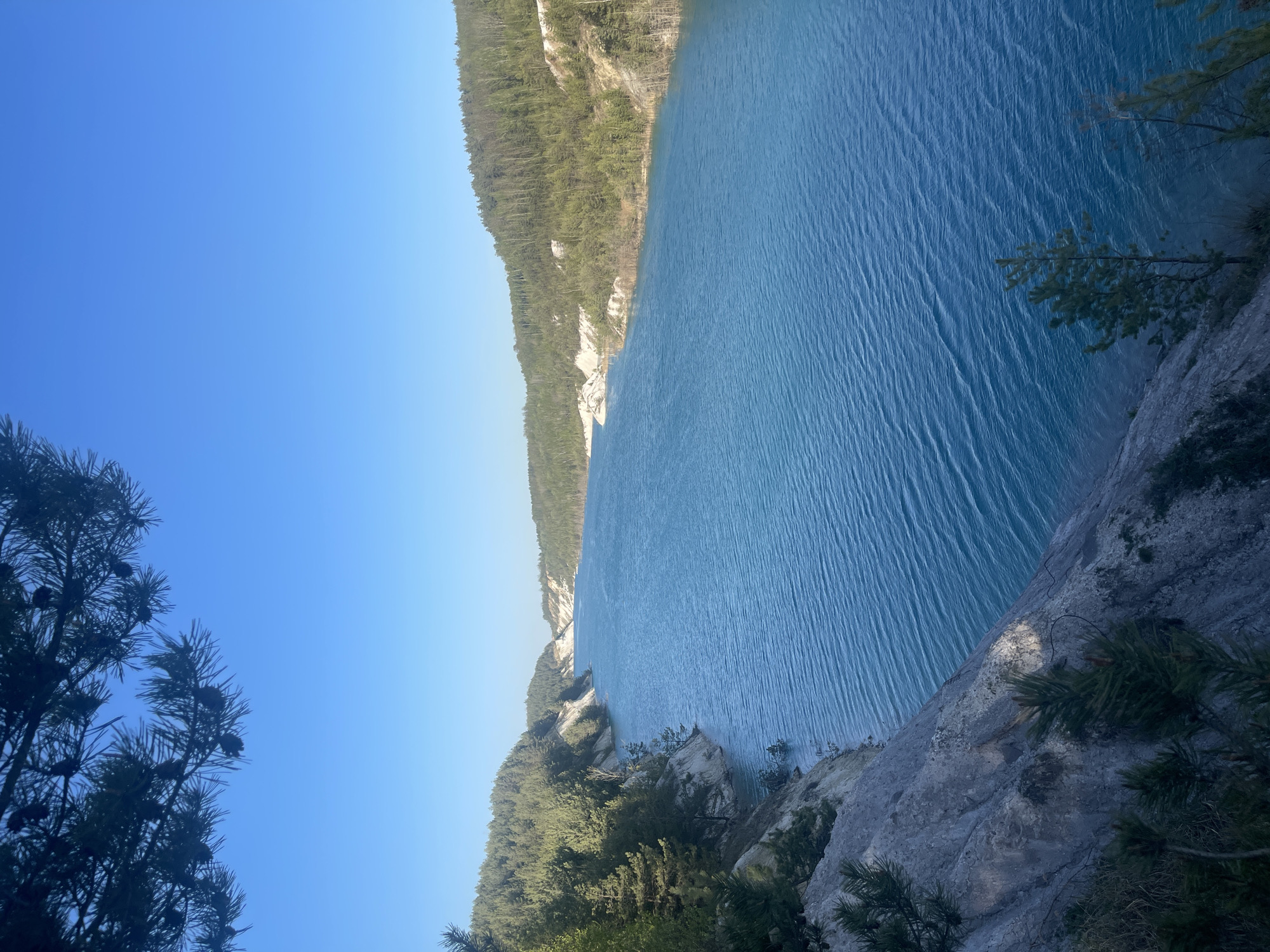 A high-angle, rotated view depicts a blue lake bordered by forested hills under a clear blue sky. The lake surface shows subtle ripples. Trees and rocky outcrops frame the bottom right and bottom left corners, creating a sense of height and perspective. The image captures a natural landscape. The location is within Краснасельскі сельскі Савет, Belarus.
