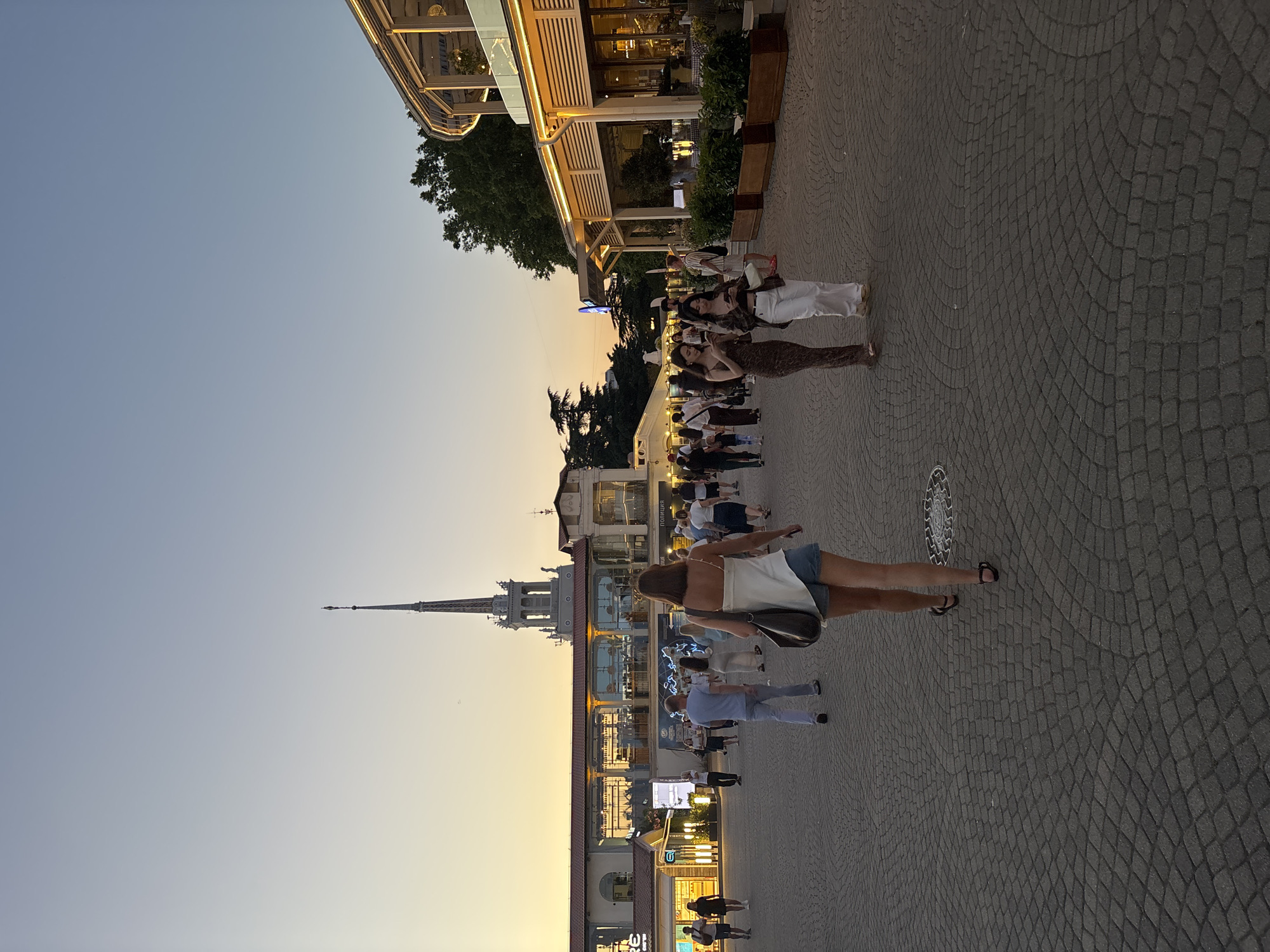 The image depicts an outdoor scene with a clear sky and warm sunset lighting, suggesting it is late afternoon or early evening. The setting appears to be a promenade or pedestrian walkway lined with buildings and shops, possibly in a coastal or resort area, given the presence of a prominent spire on one of the buildings reminiscent of maritime architecture.
Several individuals are visible, walking along the paved pathway. The foreground features a woman from the back, wearing a white top and denim shorts, carrying a shoulder bag. She is walking on the cobblestone-like pavement. Further in the background, other people are scattered, some walking in groups, others individually. Their attire suggests casual summer clothing.
The buildings have storefronts with lighting, indicating businesses are open. One building has an awning with lights, and plants are visible in planters. The overall atmosphere is relaxed and leisurely, with people enjoying an evening stroll. The sky is a gradient of soft blue and orange, typical of a sunset.
A spire, which is a dominant architectural element in the mid-ground, is a significant landmark. The pavement is made of small, dark gray stones arranged in a pattern. There is a circular manhole cover on the pavement. No discernible text is clearly visible in the image. The general ambiance suggests a pleasant, active urban environment during a beautiful time of day.