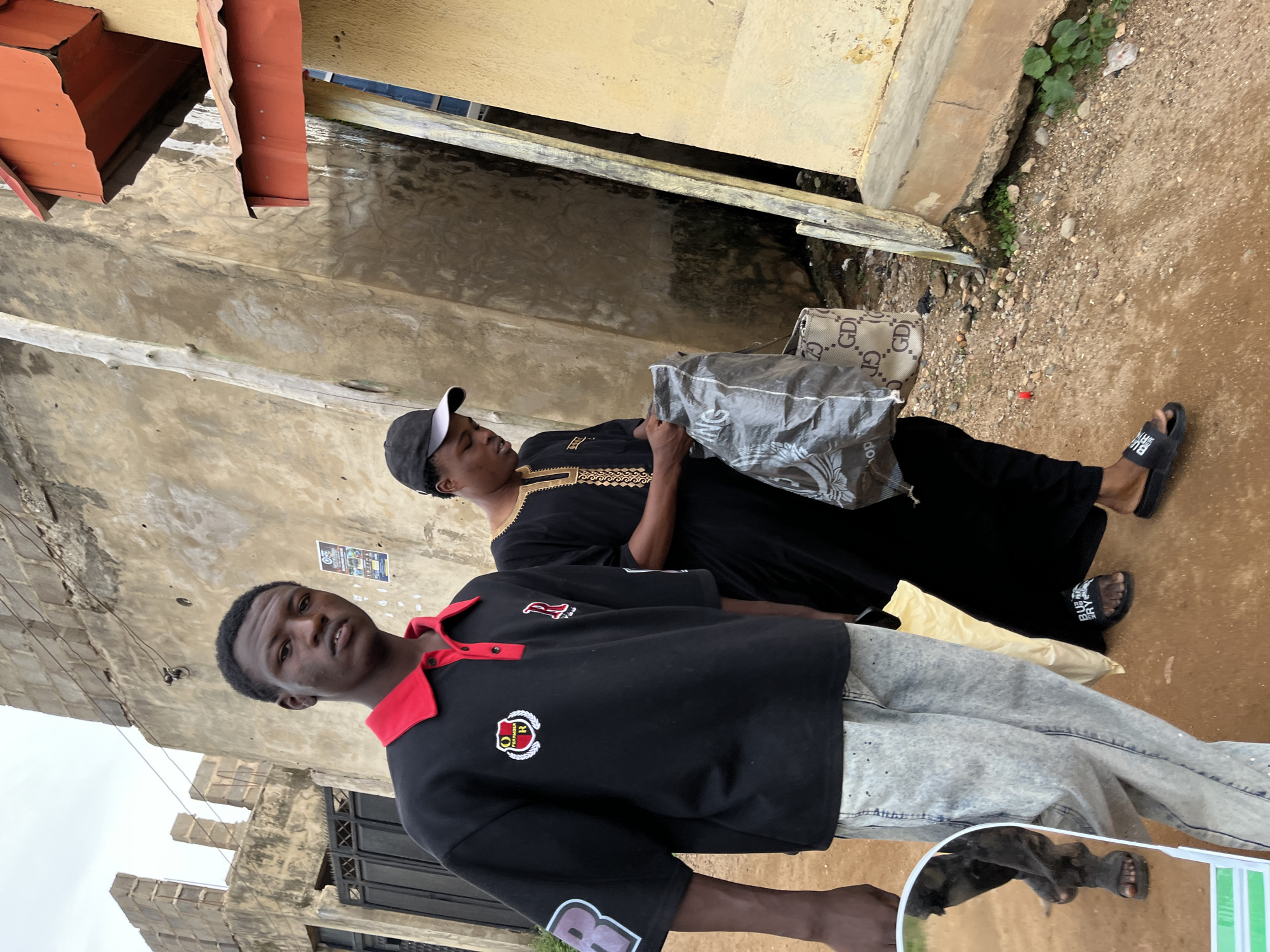 Two young men are walking down a dirt path in front of weathered, light-colored buildings. The man in the foreground, on the left, wears a black polo shirt with a red collar and trim, and light-colored, speckled jeans. He is looking at the camera. The second man, to the right, wears a black, traditional-style garment with gold trim and a black baseball cap. He is looking down and holding a large, translucent plastic bag that contains another patterned bag with "GD" logos. A bright yellow bag is visible on the ground near the second man's feet. The environment appears to be a somewhat neglected urban area, possibly in Rafin Par, Nigeria, with debris on the ground and rough building textures. A portion of a reflective surface, possibly a mirror from a vehicle, is visible in the lower-left corner, reflecting the legs of a third person wearing dark clothing and sandals.