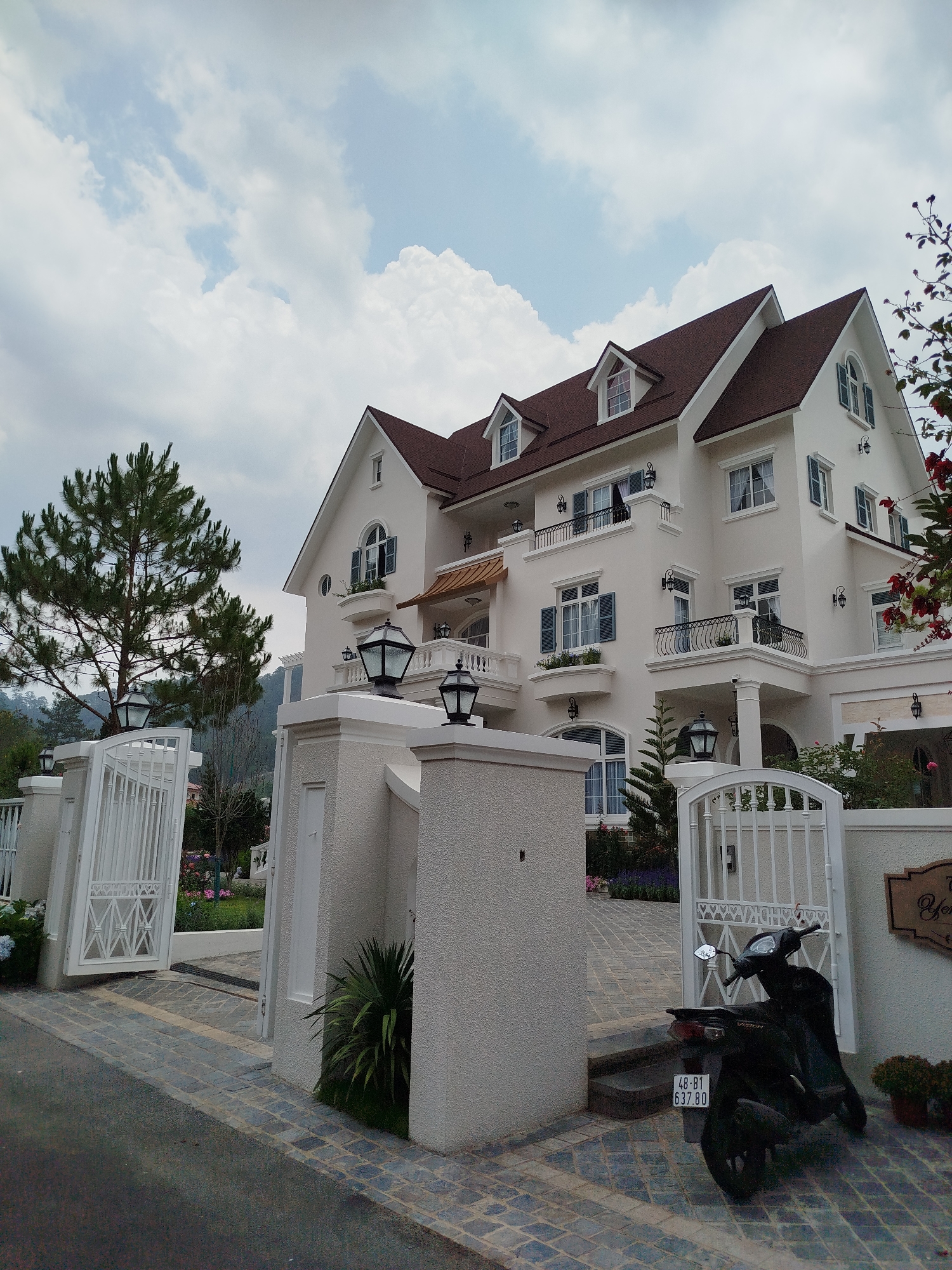 The scene depicts a large, multi-story cream-colored house with a brown roof in what is presumed to be Hiep Thanh, Vietnam. The house features multiple dormer windows, balconies with wrought iron railings, and various exterior light fixtures. The house is surrounded by a gated entrance consisting of white pillars and metal gates. A black scooter is parked near the gate. The sky is overcast with scattered clouds. A large pine tree is visible to the left of the house. The street leading to the house is paved with stone blocks. Foliage and flowers are visible near the entrance and along the house's perimeter.