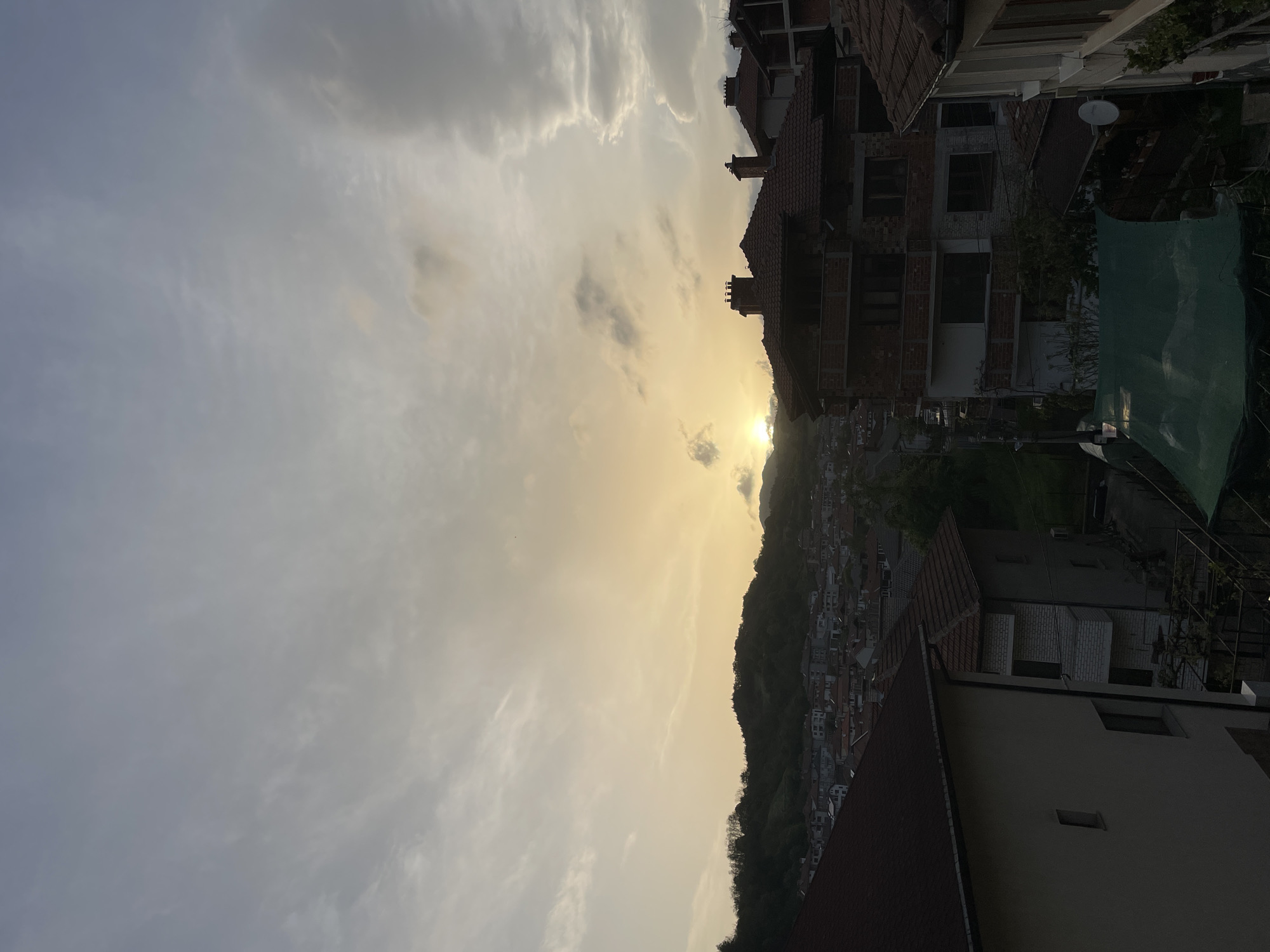 The image depicts a view over the city of Zlatograd, Bulgaria, at sunset. The sky is filled with clouds illuminated by the setting sun. A mountain ridge is silhouetted in the center of the frame, with buildings of Zlatograd extending up its side. In the foreground, there are buildings with red tiled roofs and brick or white walls. The image appears to have been taken from an elevated position, possibly from the roof or upper floor of a building.