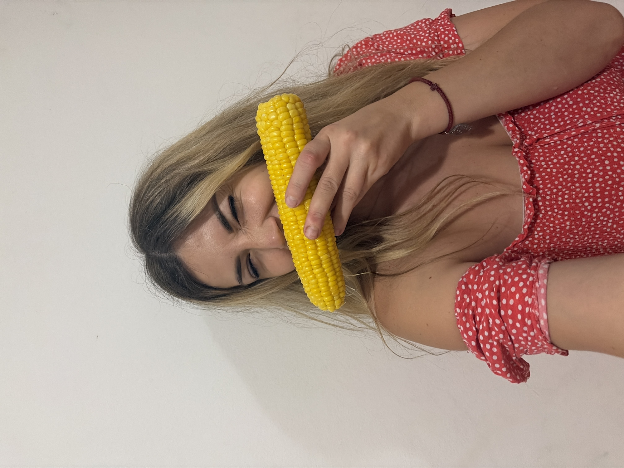 A woman with blonde hair and a red and white polka dot dress is playfully holding a corn on the cob to her mouth. She is looking at the camera with her eyes closed and a smile on her face. The background is a plain white wall, suggesting an indoor setting. The image does not provide any information about the location in Budva, Montenegro.