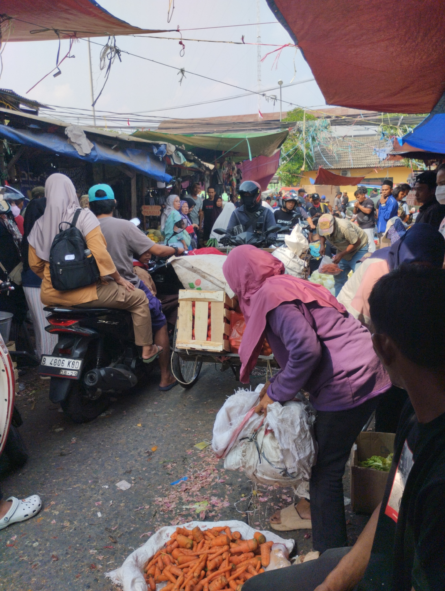 This is a medium shot from a street market in Bekasi, Indonesia. The scene is crowded with people, many of whom are wearing traditional Islamic clothing, such as hijabs. The market is covered by makeshift awnings made of tarpaulins and fabric, providing shade from the sun. Various goods, including vegetables and what appear to be fruits, are displayed in crates and bags. A motorbike with a trailer attached is visible, loaded with produce. The ground is paved and appears somewhat dirty, with debris scattered around. The overall atmosphere suggests a busy and bustling marketplace.