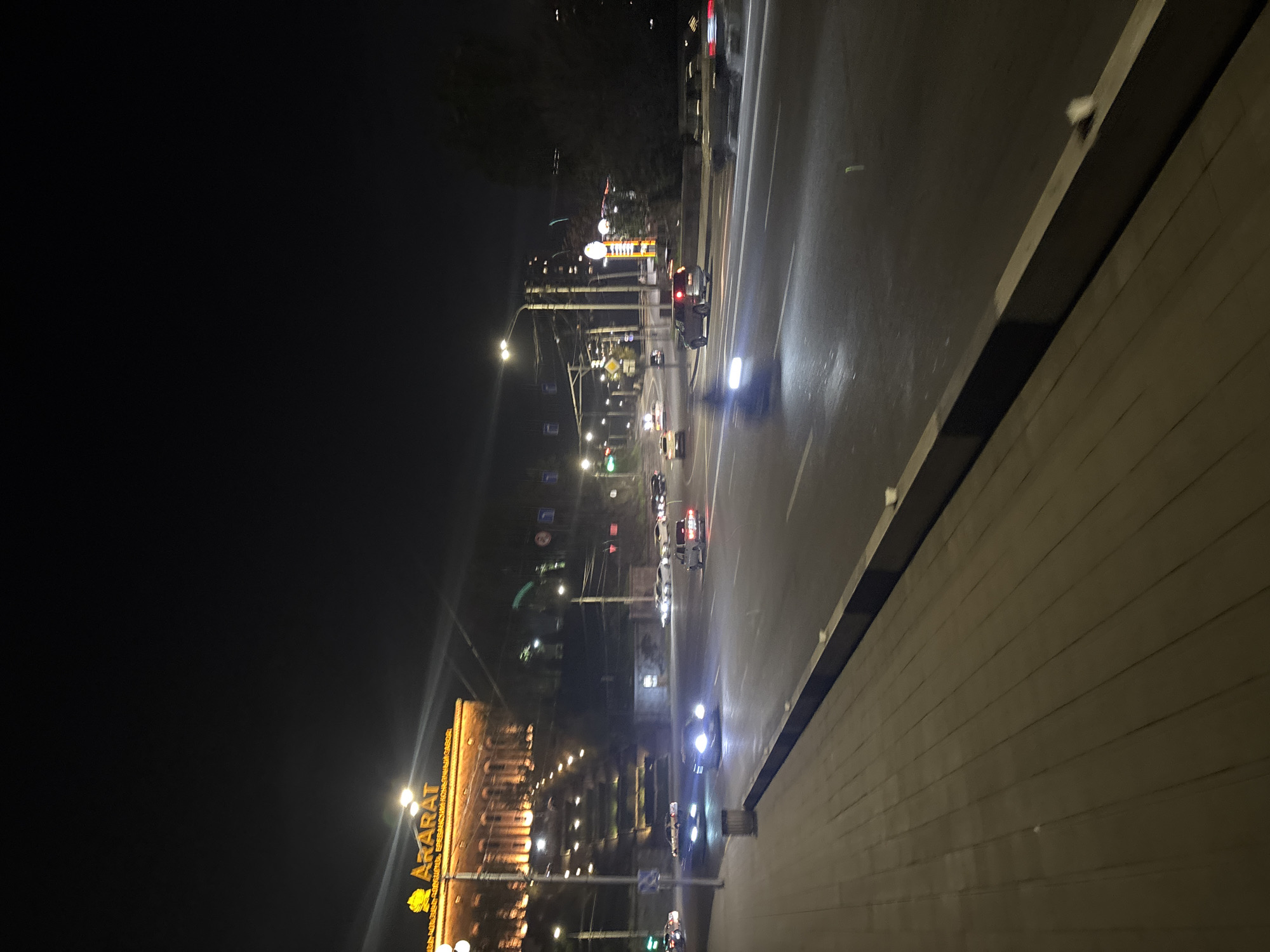 The image captures a nighttime street scene in Yerevan, Armenia. The view includes a brightly lit building with the name "ARARAT" prominently displayed, likely referring to the Ararat Brandy Factory. The image shows a busy street with cars moving in both directions, their headlights illuminating the wet pavement. Streetlights line the avenue, and the background includes other buildings and signs. The foreground features a paved walkway or sidewalk area.