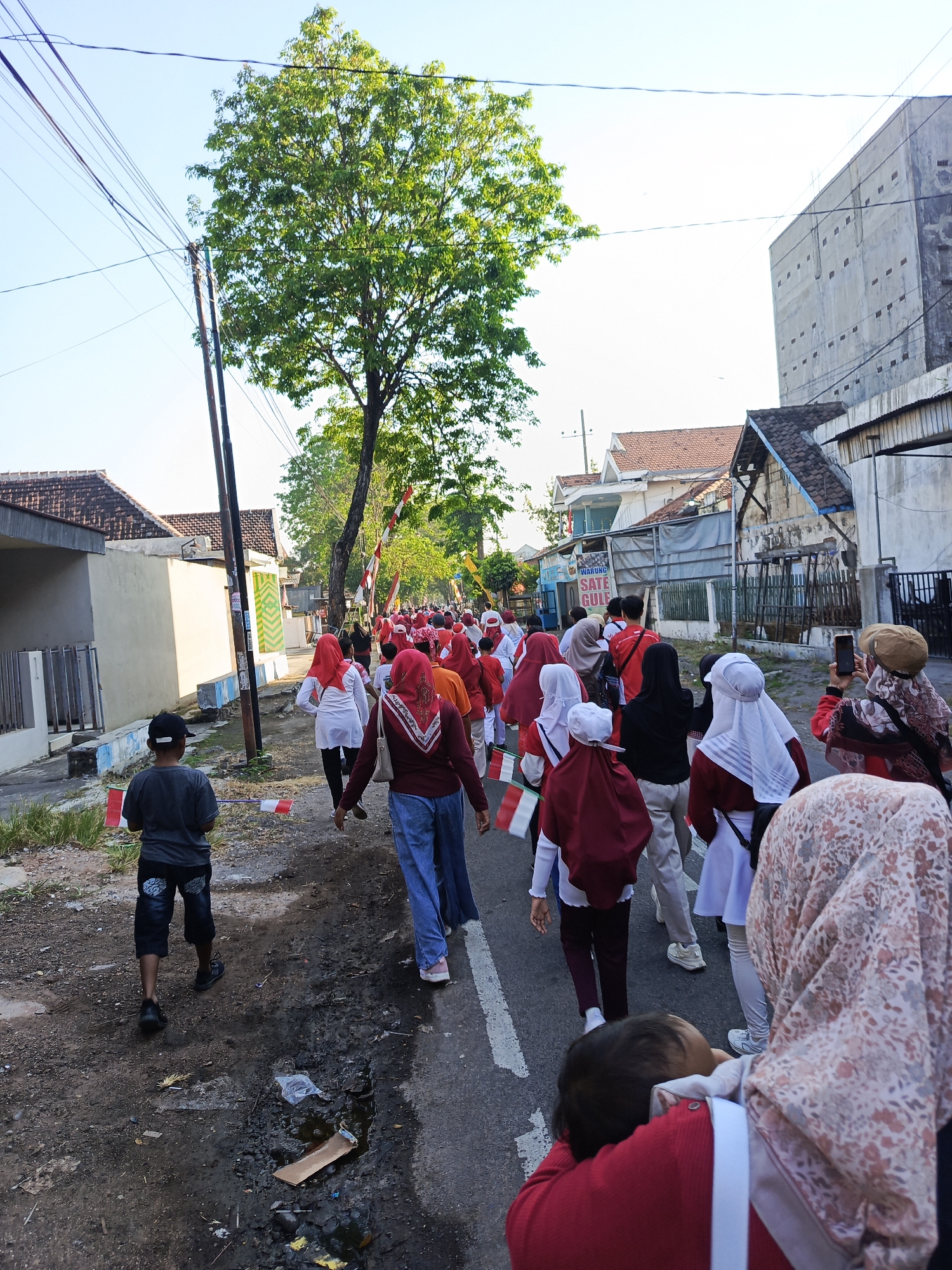 In the city of Semampir, Indonesia, a group of people, many wearing red and white attire, are participating in a procession along a paved street. The procession appears to be moving from right to left in the frame. A young boy in the foreground, facing away from the camera, holds a small Indonesian flag. Other individuals in the procession are also holding flags. Buildings line both sides of the street, some appearing residential with tiled roofs, while others are more industrial or commercial. One building in the background has a sign that reads "WARUNG SATE GULI". Electrical wires and poles are visible overhead. The street surface shows signs of wear, with some potholes and debris. The sky is clear and blue, suggesting daytime.