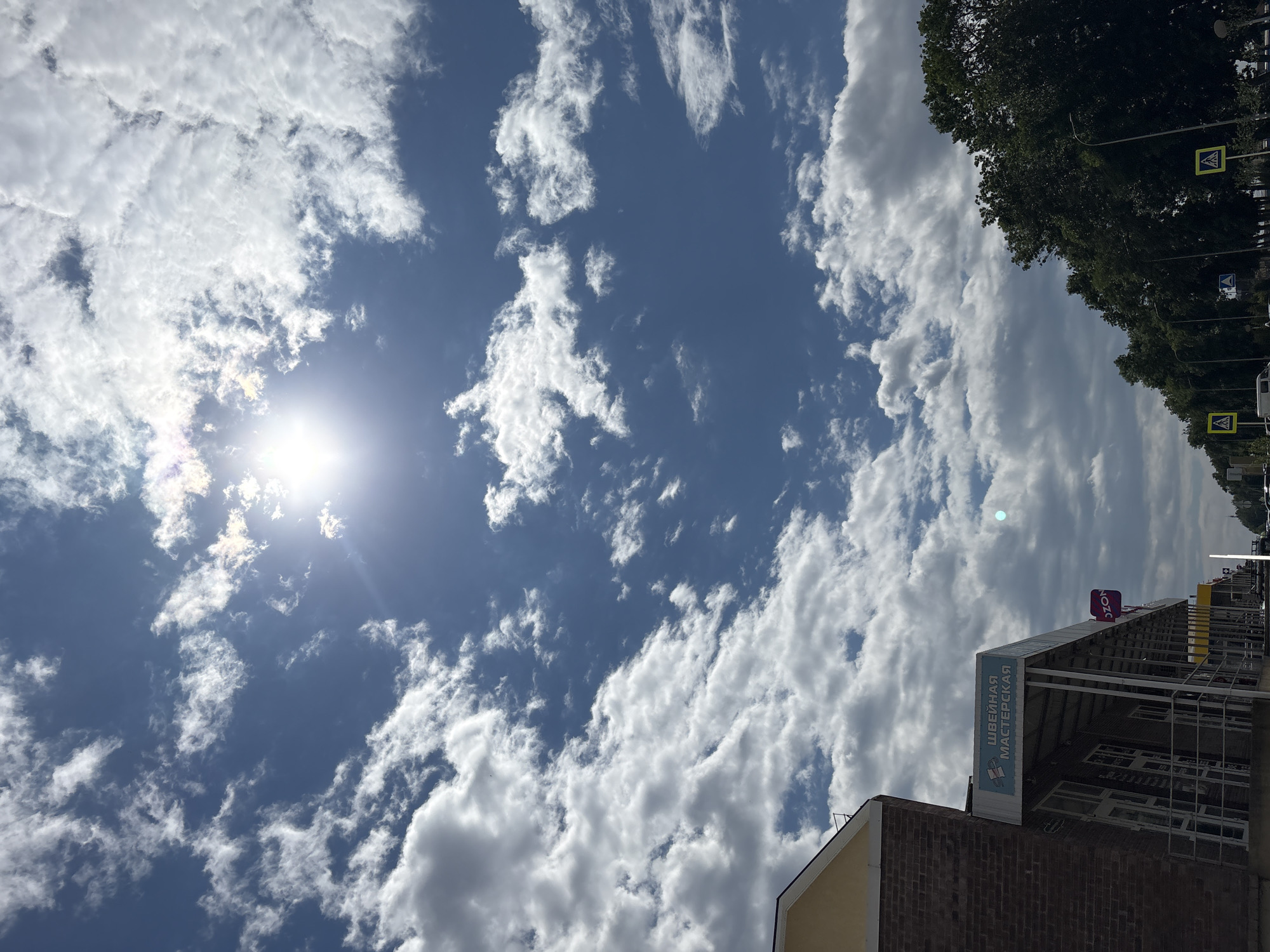 This is a low-angle, vertical, outdoor, daytime photograph of a street scene in Mikhaylovsk, Russia. The sky is a vibrant blue, filled with scattered white cumulus clouds. The sun is visible near the top center of the frame, shining brightly and creating a lens flare effect. In the foreground, on the left side of the image, is the corner of a brick building with a light yellow facade and a white awning. A sign on the awning reads "ШВЕЙНАЯ МАСТЕРСКАЯ" (Sewing Workshop) in blue lettering. Below the awning, there are storefronts with large glass windows. To the right of the building, a street is visible, lined with dark green trees. Several streetlights and yellow pedestrian crossing signs with black arrows are present along the street. A dark-colored vehicle is partially visible in the distance. The overall impression is a sunny day in a town.