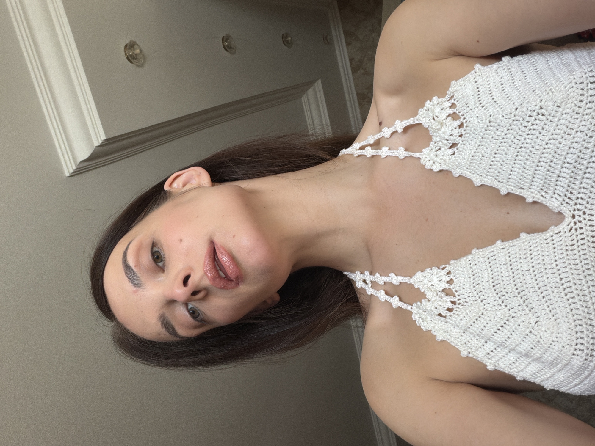 The image shows a close-up, upward-angle shot of a woman's face and upper body. She has long, dark brown hair, fair skin, and is wearing a white crocheted halter top. Her eyes are a light green, and her lips are painted with a shimmery, neutral-toned lipstick. She is looking slightly away from the camera, with her head tilted. The background is out of focus, showing a neutral-colored wall and a section of decorative ceiling molding. There are two visible light fixtures on the ceiling. The image does not provide any information about the location being Ufa, Russia, nor any other geographical context. The scene is indoors, in what appears to be a room with white-painted walls and ceiling.