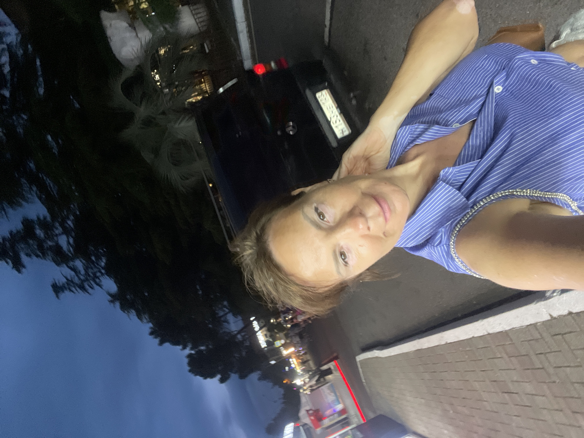 A woman, likely in her 50s, takes a selfie outdoors at dusk, possibly in the Khostinskiy district of Sochi, Russia. The background reveals a street scene with a dark blue sky, silhouetted trees, and a dark colored van with a license plate. Lights from businesses and street lamps illuminate the street, which is paved with bricks and bordered by a curb. The woman is wearing a blue and white striped sleeveless shirt with silver embellishments on the shoulders. Her right hand is touching the back of her head. The overall lighting suggests it's twilight.