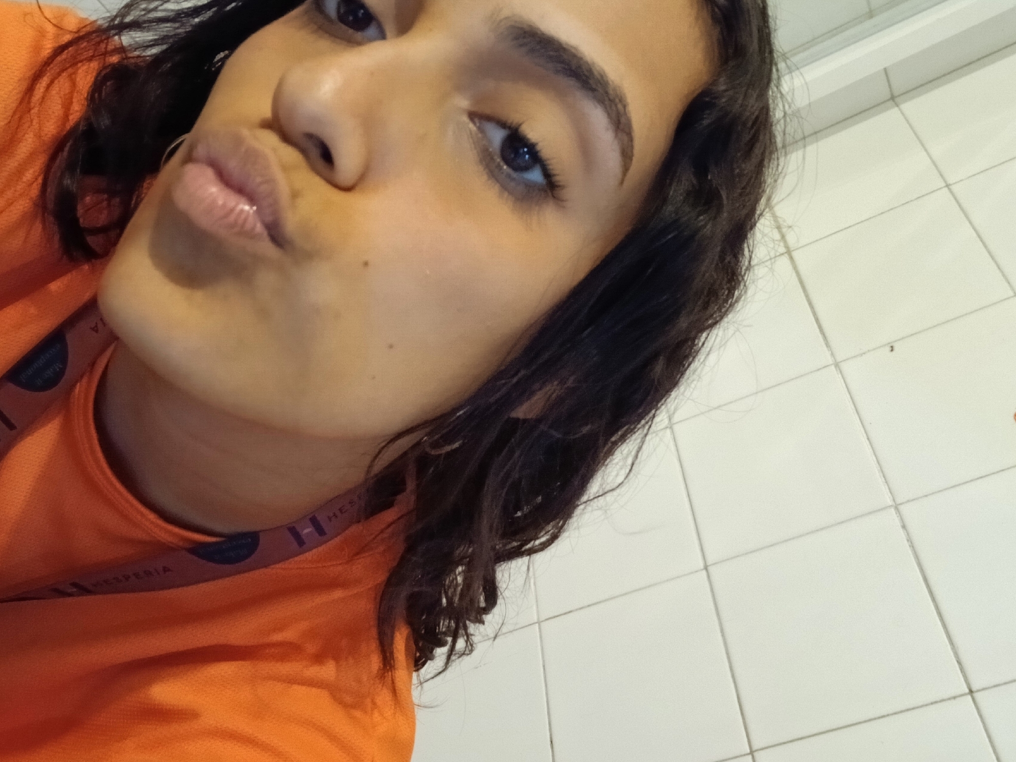 A close-up, tilted selfie captures a young woman with dark, wavy hair and dark eyes. She is making a playful "kiss face" with her lips puckered. Her complexion is warm-toned, and her eyebrows are well-defined. She is wearing a bright orange top, which appears to be part of a uniform or work attire. Around her neck, a lanyard or strap is visible, featuring the text "HESPERIA" repeated multiple times, along with an "H" logo. Smaller, partially visible text on the strap also reads "Made it".
The background consists of light-colored, possibly white, square tiles with dark grout lines, suggesting an indoor setting such as a wall in a bathroom or service area. The lighting appears to be indoor, creating soft shadows. The general location context for the image is Playa El Agua, Venezuela.