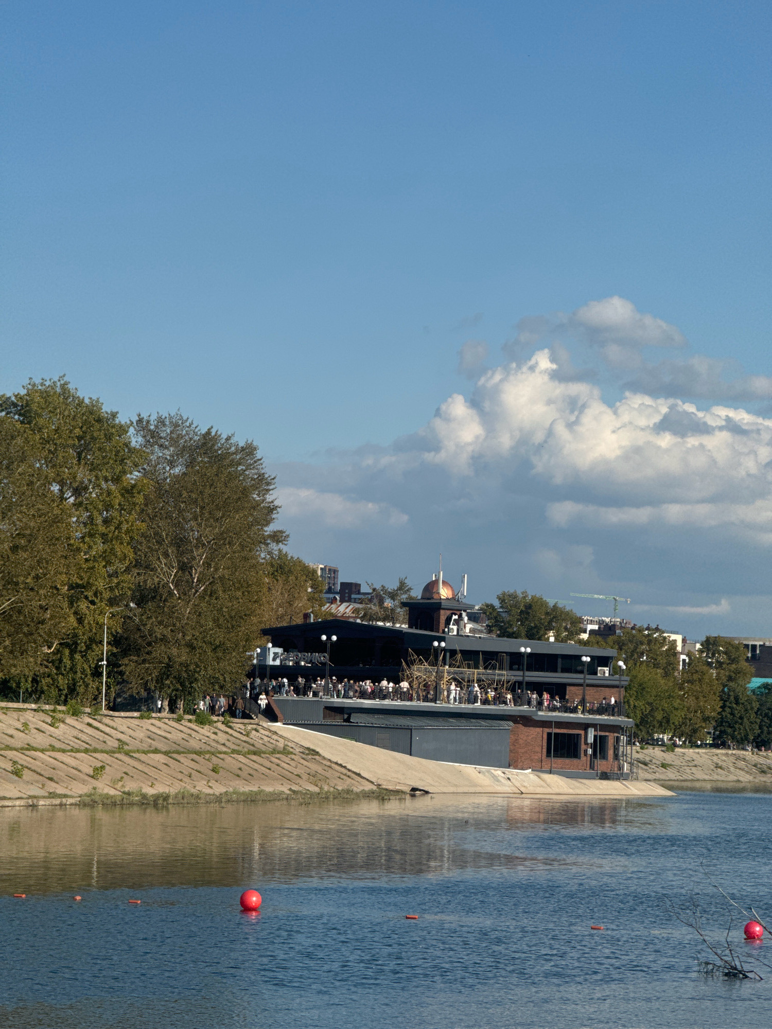 A wide-angle, eye-level shot captures a lively scene along a riverbank in Irkutsk, Russia. The foreground is dominated by a wide, blue body of water reflecting the bright, partly cloudy sky. Several orange buoys are visible in the water, with a large red buoy to the right of center.
The midground features a tiered, modern building situated on the riverbank, possibly a restaurant or entertainment venue. People are gathered on its upper levels and along its exterior, suggesting a social gathering or event. The building is a mix of dark siding and red brick accents, with prominent lighting fixtures. Behind the building, a line of trees with lush green foliage runs along the riverbank, interspersed with several urban structures, including buildings and what appears to be a construction crane in the distance.
The riverbank itself is a sloping concrete structure, with diagonal lines creating a terraced effect. Some vegetation is visible growing in the crevices of the concrete. The overall atmosphere is one of a pleasant day, with many people enjoying the riverside environment. The city skyline is visible in the background, suggesting an urban setting.