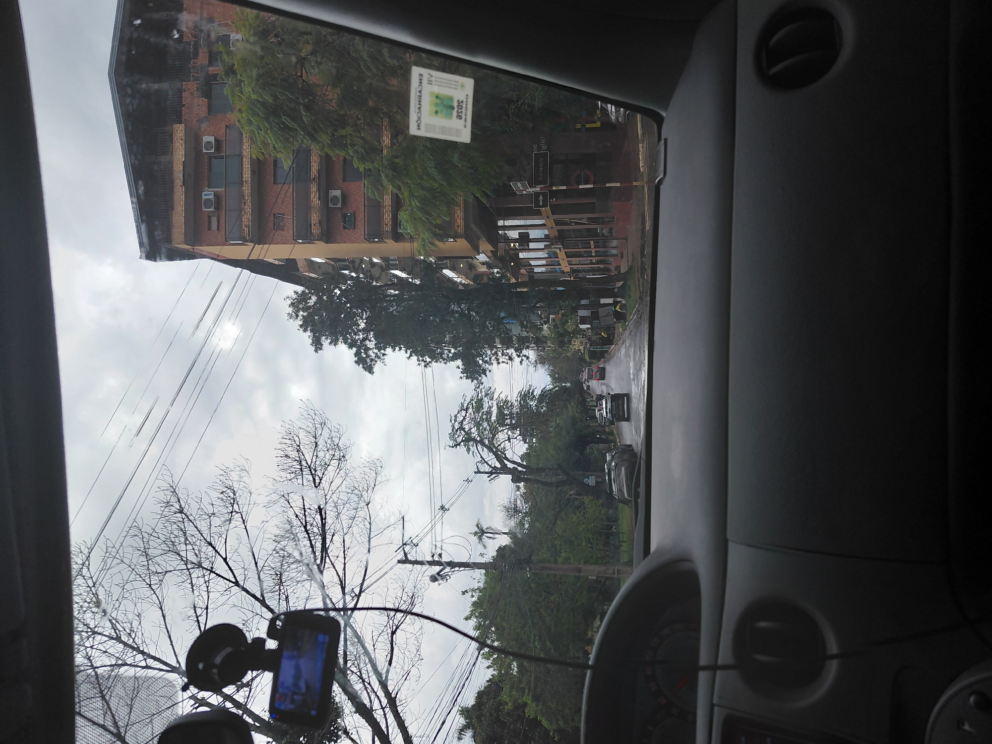 The image is taken from inside a vehicle, looking forward through the windshield. The view encompasses a street scene in Encarnacion, Paraguay, under an overcast sky. In the foreground, the dashboard of the car is visible, with a portion of the instrument cluster showing speed markings. A dashcam is mounted on the windshield, displaying a blue screen.
The street outside is wet, suggesting recent rain. Cars are driving on the road, including a dark sedan in the middle ground and a red bus further down. On the right side of the street, a multi-story brick building with balconies and air conditioning units is prominent. Trees with sparse foliage line the street, and utility poles with numerous wires are visible. On the building's window, a white sticker with text and a barcode is affixed. A street sign indicating "European English" with an arrow pointing right is also visible. The overall atmosphere is urban and somewhat somber due to the cloudy weather.