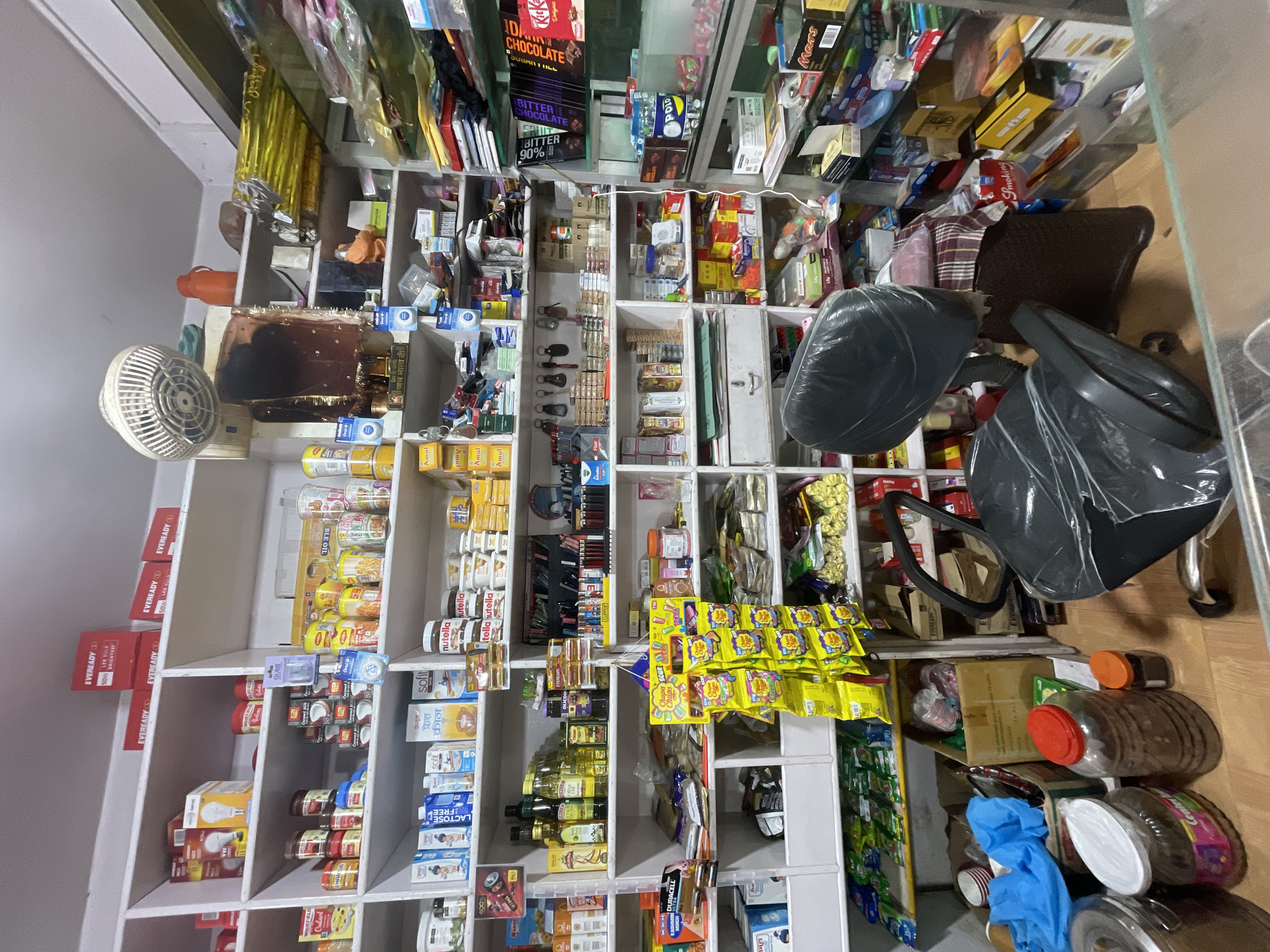 The image shows the interior of a small, cluttered convenience store in Bhagsu, India. Shelves are packed with a wide variety of items including packaged food products, toiletries, household supplies, and electronic goods. Some prominent items include boxes of Eveready batteries and bulbs, jars of what appear to be pickles or preserves, bottles of oil, and several boxes of Nutella. There are also bags of chips, candy, and various other snacks. In the foreground, there are two office chairs, both covered in plastic sheeting. One chair is positioned in front of a counter that is partially visible. The overall impression is one of a busy, well-stocked shop with merchandise stacked high.