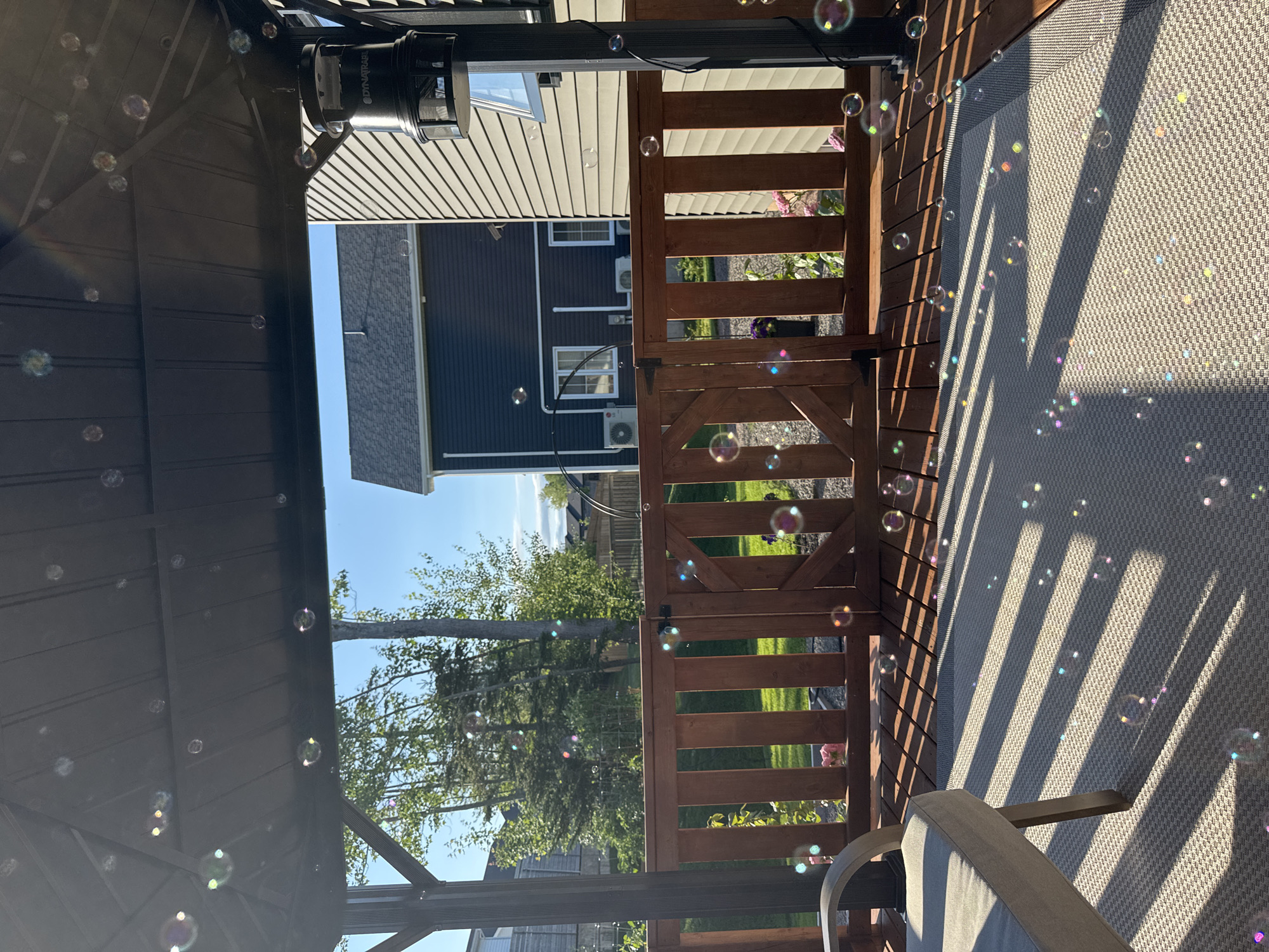 The image depicts a backyard deck area in Dieppe, Canada, under a gazebo. The scene is filled with numerous soap bubbles reflecting sunlight. A grey outdoor chair is positioned on the left side of the frame, situated on a patterned rug. The deck itself is made of wooden planks, and a wooden fence with a gate encloses the area. Beyond the fence, a grassy lawn and trees are visible, along with residential houses under a clear blue sky. A bug zapper device, labeled "DYNATRAP", is mounted on a black pole on the right side of the gazebo. The overall impression is a sunny, outdoor recreational space.