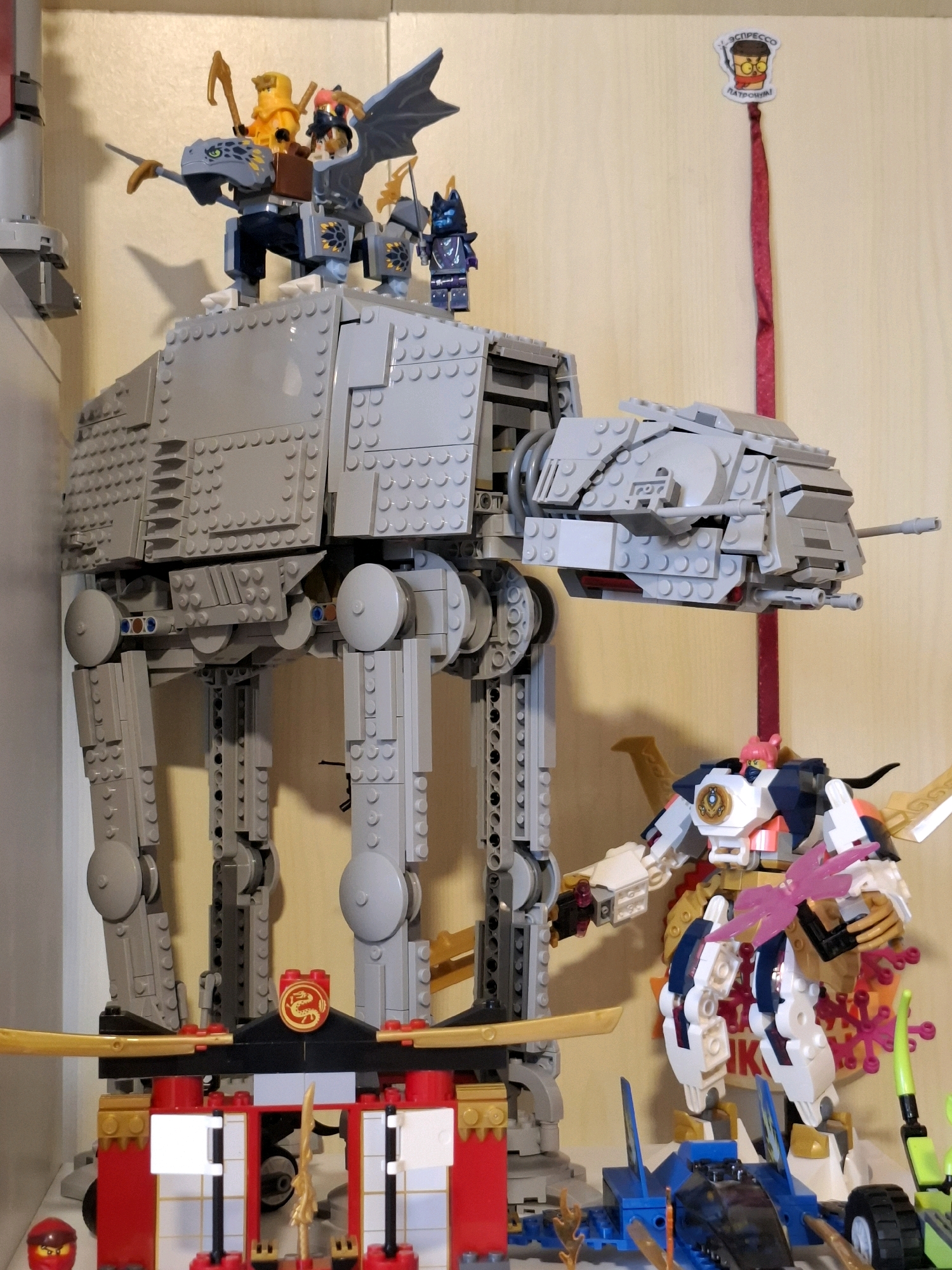 The image shows a collection of LEGO toys displayed on shelves. A large gray AT-AT Walker toy from Star Wars is the central focus, with several minifigures and a LEGO dragon perched on top of it. To the right of the AT-AT, a large white and blue mecha-style LEGO robot is positioned. In front of these, a red and gold LEGO structure resembling a temple or gate is visible, with a smaller red LEGO ninja figure at the bottom left. A blue LEGO vehicle is also present in the foreground, to the right of the temple. The background is a plain, light-colored wall. There is no indication of the city of Tyumen, Russia, or any real-world urban environment. The scene is entirely composed of toy models.