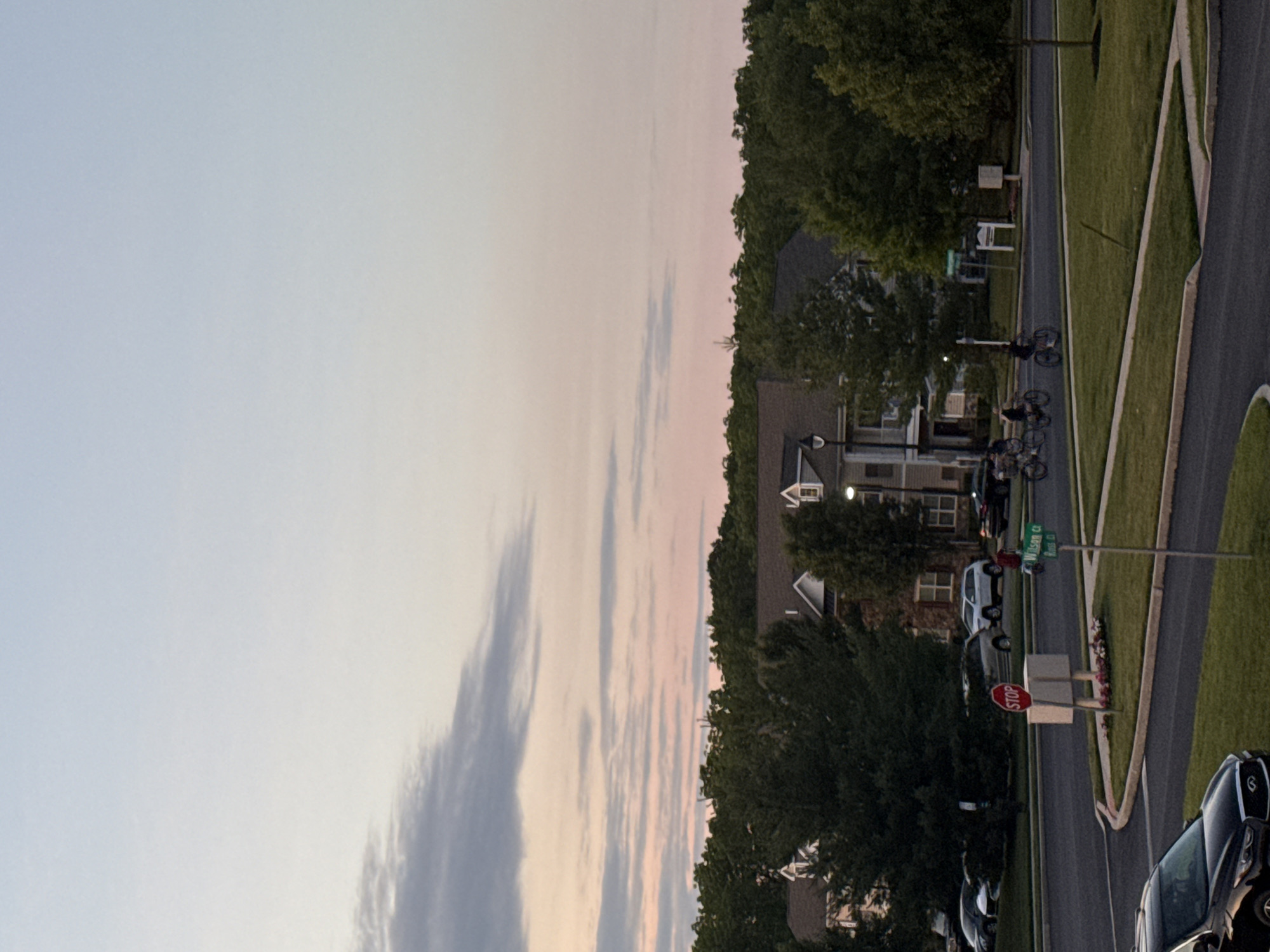 The image captures a suburban street scene in Allen Township, United States of America, during a twilight hour, with a pale blue and pink gradient sky. A line of trees borders the horizon, behind which residential buildings with peaked roofs are visible. In the foreground, a dark-colored sedan is parked on the left side of the frame. A road curves through the scene, with a stop sign positioned on a traffic island in the middle of the intersection. A green street sign indicates "Wilson Ct" and "Rose B." A group of individuals is riding bicycles on the road in the middle ground, heading towards the right of the frame. Several cars are parked in the background along the street.