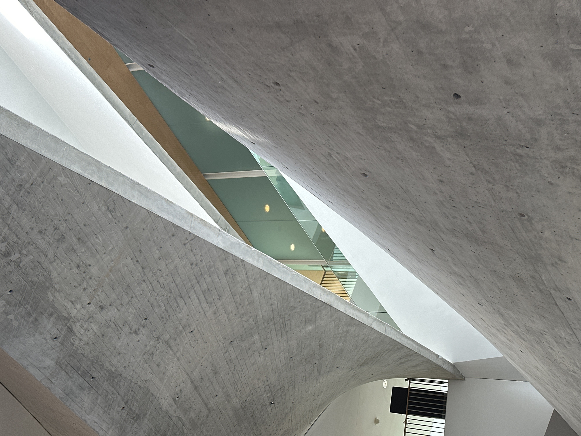 The image captures an upward view within the Tel Aviv Museum of Art in Tel Aviv, Israel, specifically focused on the architectural design of the Herta and Paul Amir Building. The composition highlights intersecting planes of cast concrete, forming a complex geometric space. Portions of the interior structure are visible, including glazing that admits natural light, creating a contrast between the raw concrete and the lighter interior spaces. The scene is devoid of people, emphasizing the structural elements and their interplay. A small section of a black screen or ventilation panel is visible in the lower right, providing a sense of scale. The overall mood is one of architectural observation and appreciation of design.