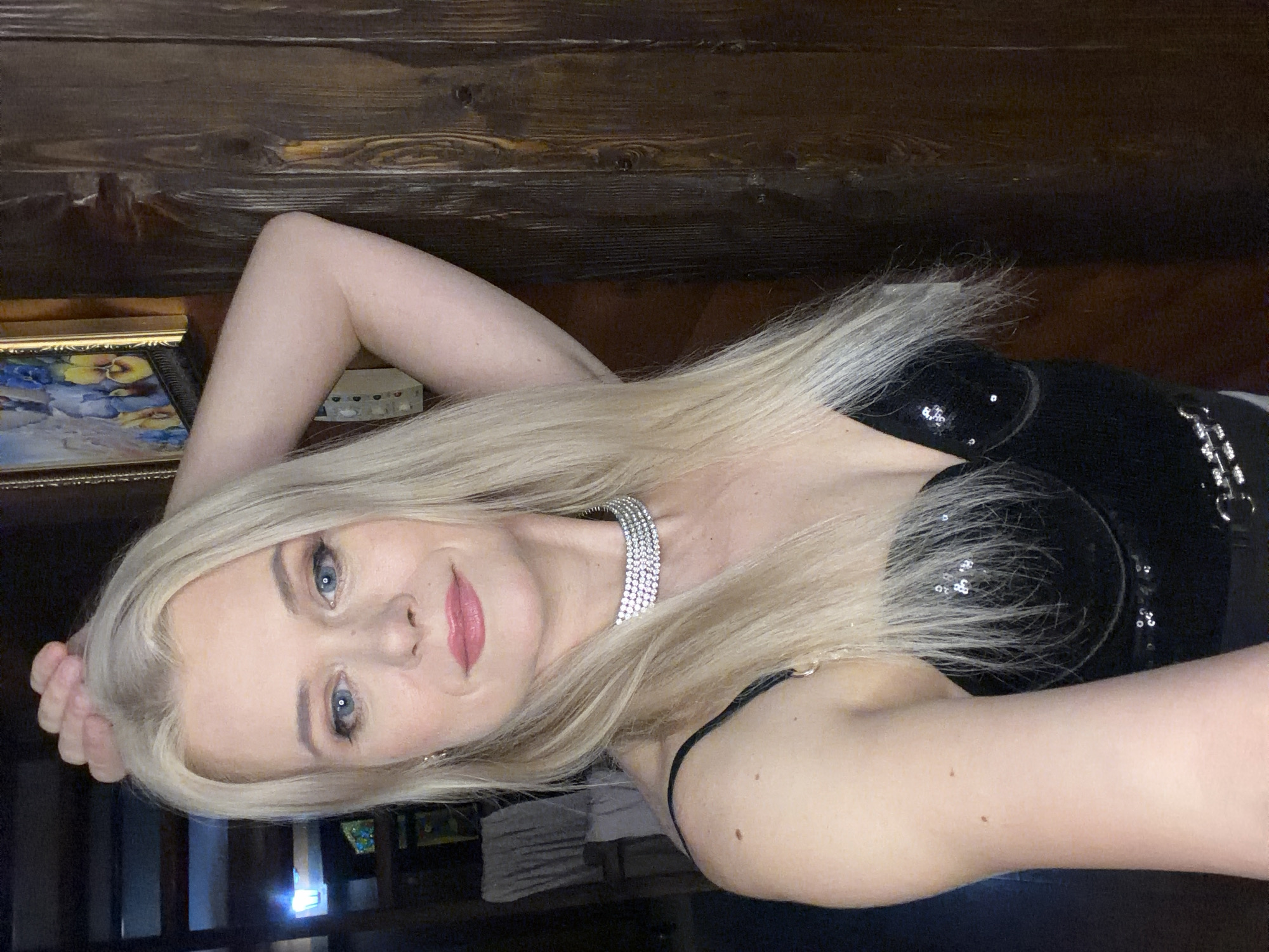 A blonde woman with blue eyes and pink lipstick is captured in a selfie, with her left arm raised to her head. She is wearing a black sequined top and a jeweled choker necklace. The background features a wooden wall and a framed picture on the left. The scene appears to be indoors, possibly in a residential setting in Sochi, Russia.