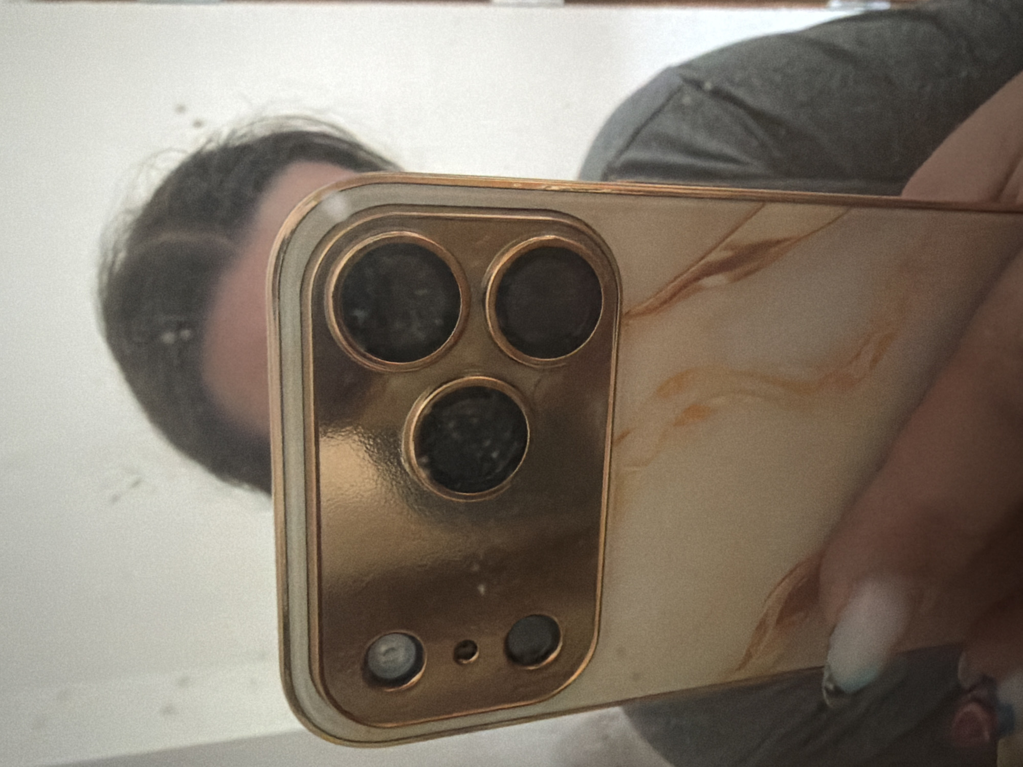 The media file shows a close-up, reflective image captured in a mirror. The primary subject is a smartphone with a distinctive camera array, held by a hand with neatly manicured nails. The phone case has a marbled pattern with golden and beige tones. The reflection in the mirror reveals a person's head and shoulder, indicating the picture is a selfie taken with the phone's rear camera.
The person's hair is dark, and they appear to be wearing a grey t-shirt or sweatshirt. The background is a plain, light-colored wall, suggesting an indoor setting. There are a few small, dark specks on the wall's surface, possibly dust or imperfections. The lighting appears to be natural, perhaps from a window, casting a soft illumination. The overall mood seems casual and everyday.
No visible text is present in the image. The reflection provides a glimpse of the person's face, but it is partially obscured by the phone and the angle of the shot, making detailed facial features difficult to discern. The focus is primarily on the phone and its camera setup, suggesting it might be a product shot or a demonstration of the phone's design. The location context of Szczecin, Poland, does not offer any specific visual cues within the image itself.