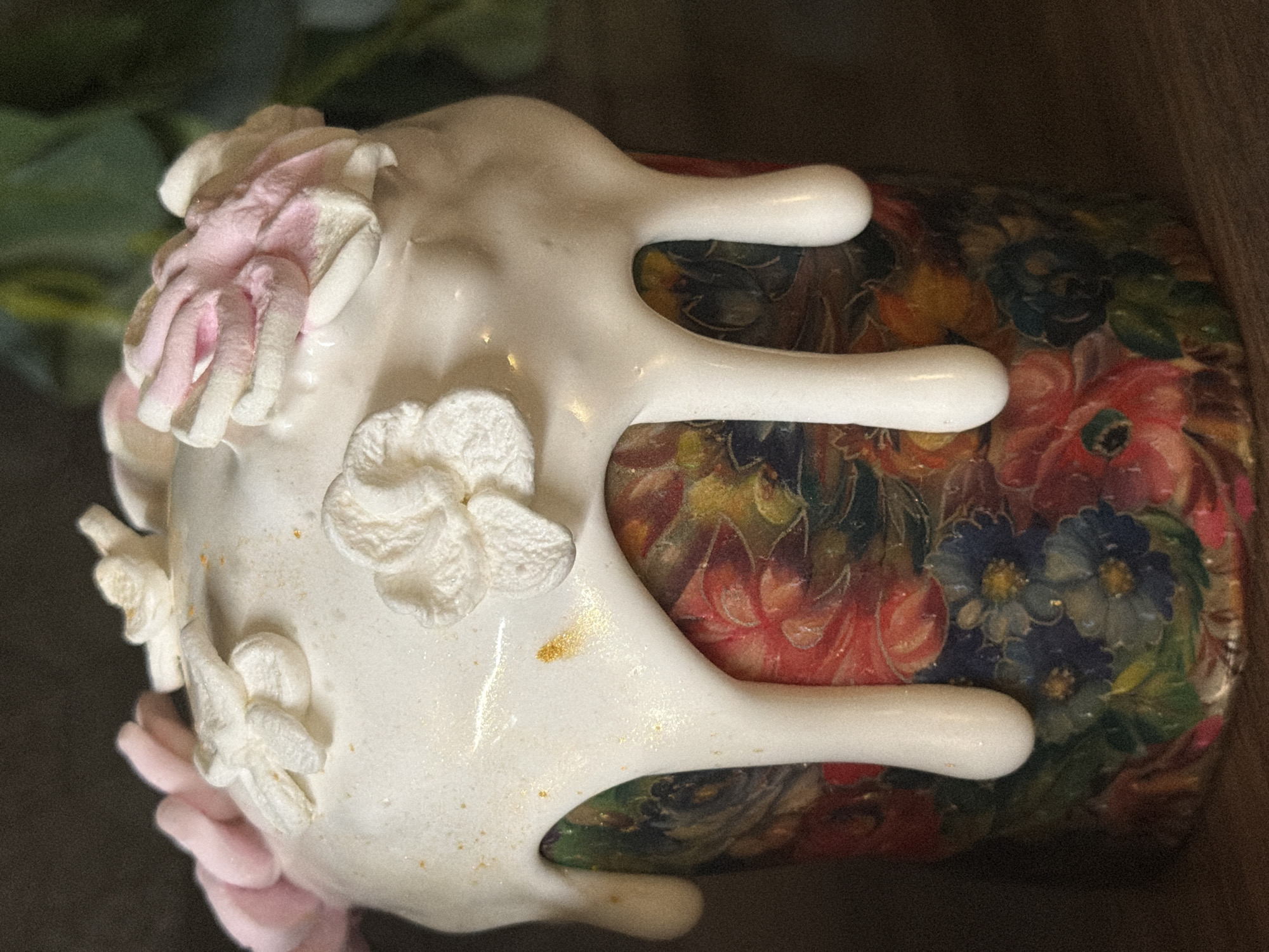 A horizontally oriented close-up shot features a ceramic object with a floral design. The object has a cylindrical body decorated with a dense pattern of flowers in various colors including red, blue, and green. Overlapping this floral design is a white, somewhat irregular, coating that appears to be a combination of smooth, glossy areas and textured floral embellishments. Some of these floral embellishments are colored in a pale pink hue. There are also small, seemingly unintentional speckles of gold color on the white coating.
The lighting is somewhat dim, casting shadows that obscure some details of the object and its environment. The object is resting on a dark surface, with a wooden background visible in the upper right corner of the image. Green foliage is out of focus in the upper left.
The location in the city of Perm, Russia is not verifiable from the provided image.