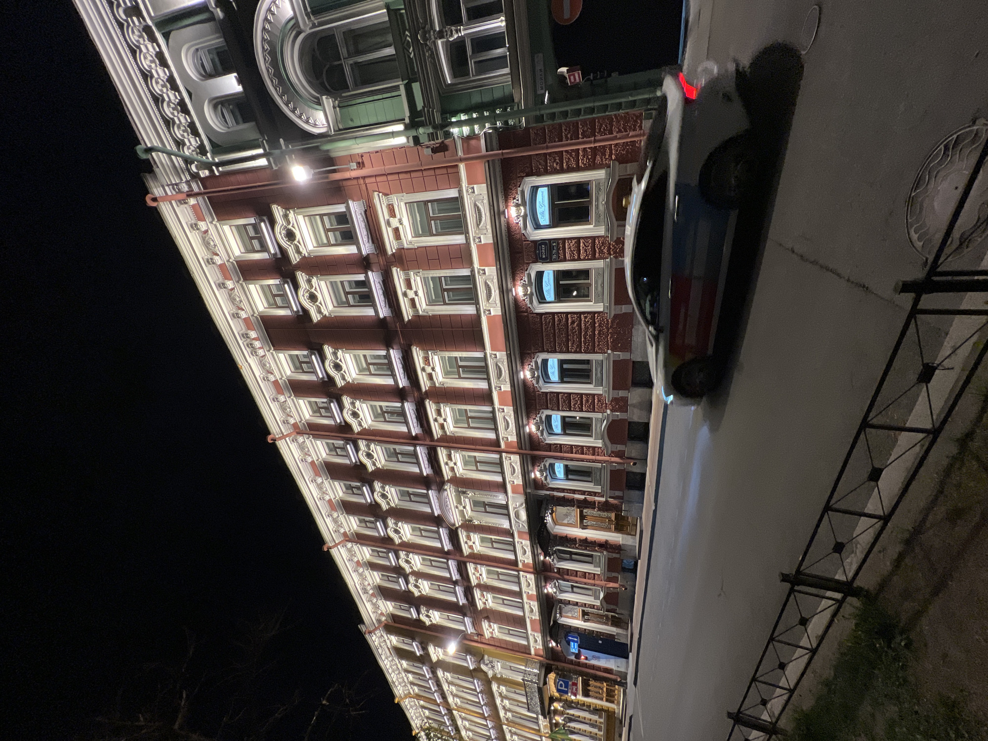 The image shows a tall building with multiple floors, likely an apartment building or hotel, located on a street in Saint Petersburg, Russia. The building's facade is decorated with white trim and ornamental details around the windows. A car is parked on the right side of the street. It is night, and the building is illuminated by street lights. A black metal fence separates the sidewalk from a small patch of greenery.