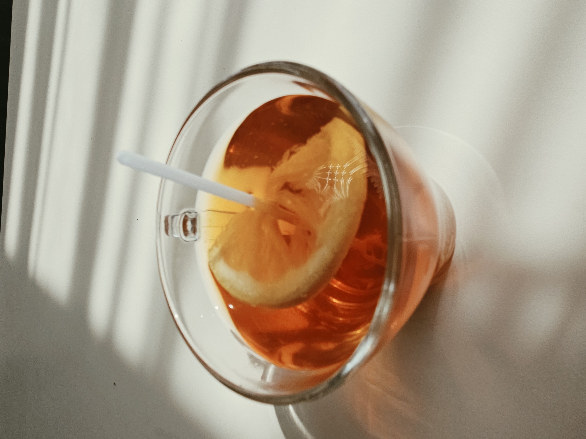 A glass mug containing an amber-colored liquid, possibly iced tea or a cocktail, sits on a light-colored surface. A slice of orange or lemon is submerged in the liquid, and a white straw is placed in the mug. Sunlight filters through blinds, casting striped shadows across the background and the mug, creating a dappled effect. The scene appears to be indoors, likely on a table or countertop. The setting is not specified to be in Ulyanovsk, Russia.