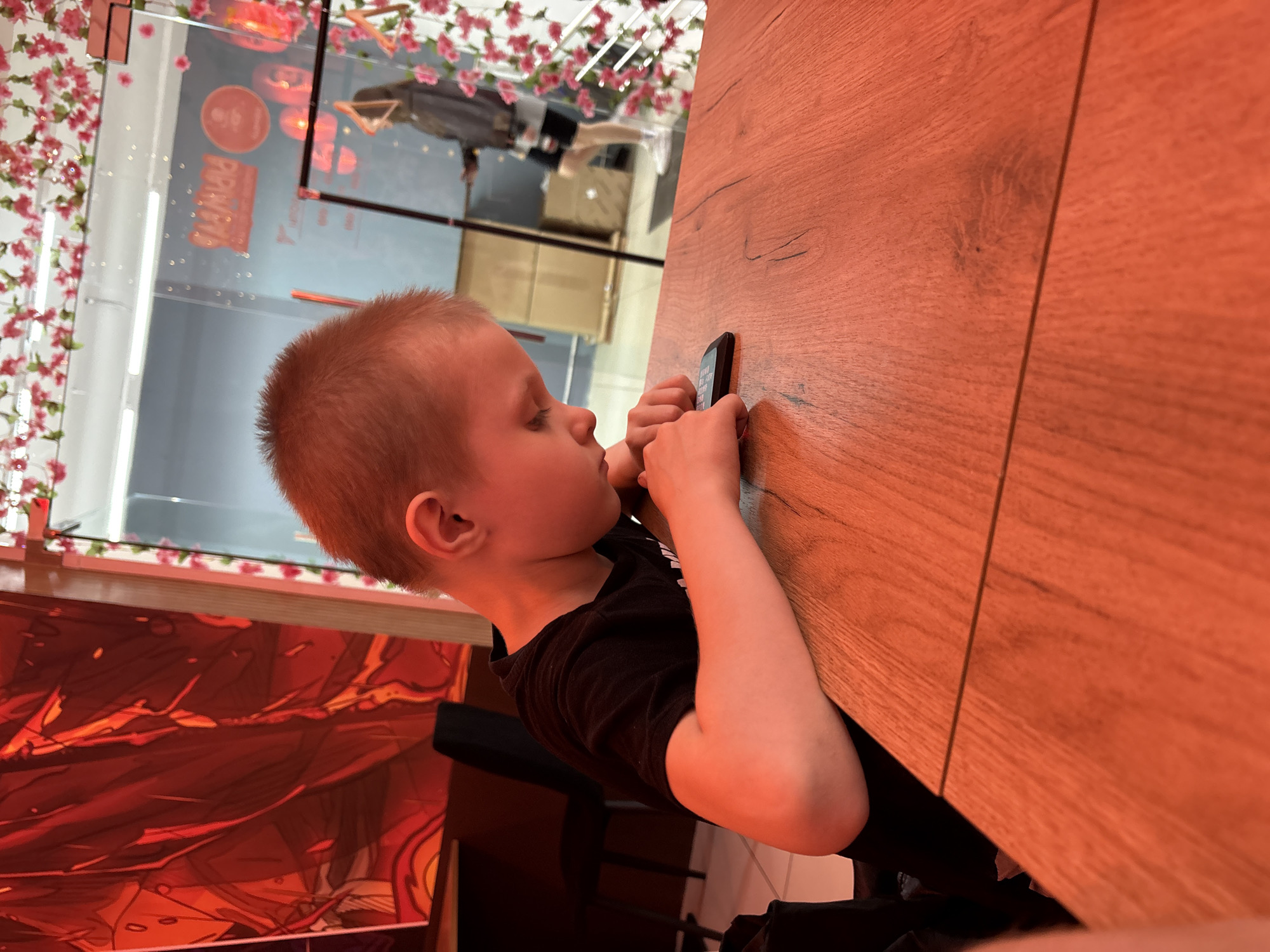 A young boy with short, light brown hair is seated at a wooden table, looking down at a black smartphone in his hands. He is wearing a black t-shirt. The table has a visible seam running horizontally across its surface. In the background, there is a large red-and-orange mural on the wall to the left. To the right, a glass partition is decorated with artificial pink flowers. Through the glass, a person in a dark jacket and shorts can be seen, as well as cardboard boxes. The lighting in the scene is warm and orange-toned. The image was taken in Saint Petersburg, Russia.