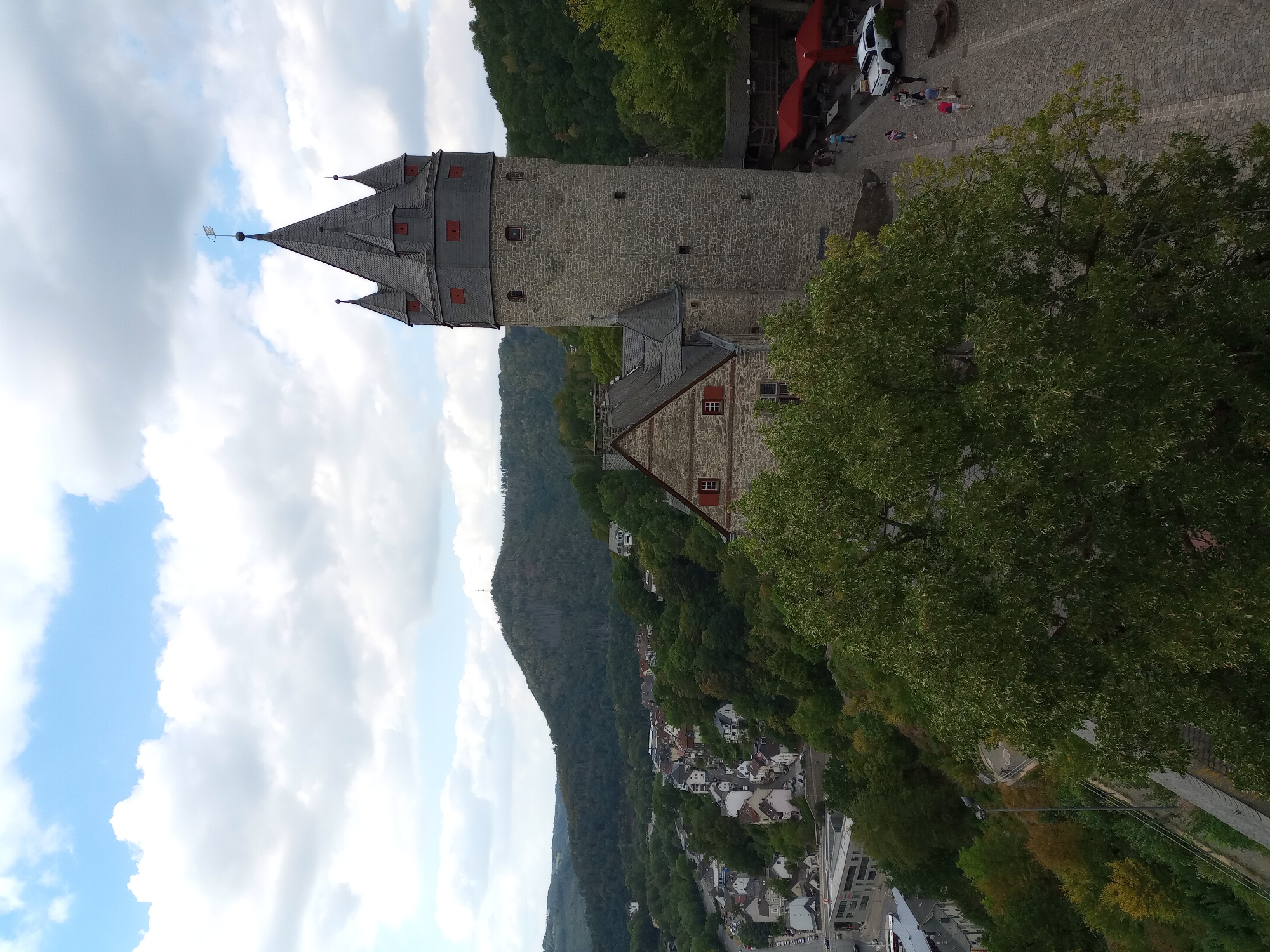 The image shows the Altena Castle (Burg Altena) in Altena, Germany. The castle is situated on a hillside overlooking the town and a river valley. The foreground features a large green tree and a paved courtyard with several people walking. A white pickup truck is parked in the courtyard, and red umbrellas cover outdoor seating areas. The castle itself is a prominent stone structure with a tall, round tower topped with a dark, multi-gabled roof. Adjacent to the tower is a section of the castle with a stone facade and a red-tiled roof. In the background, the town of Altena is visible with its houses nestled amongst lush green trees and rolling hills. The sky is partly cloudy with a bright blue sky visible.
