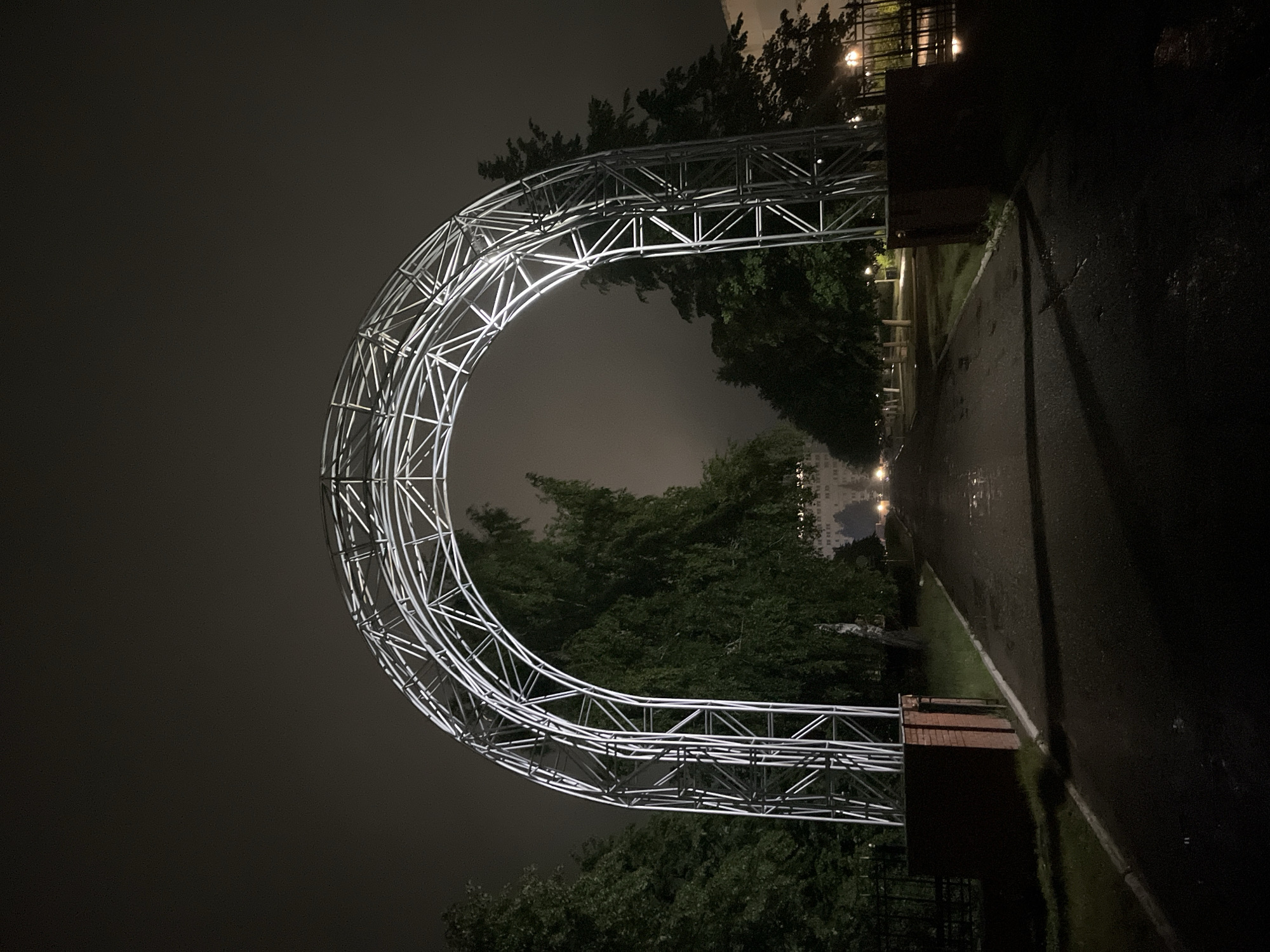The image depicts a nighttime scene in Chaikovsky, Russia, centered on a large, illuminated metal archway spanning an asphalt road. The archway is constructed from a white, latticed truss structure, emitting a bright white light that defines its form against the dark, overcast, and hazy sky. Its base rests on two reddish-brown brick pillars positioned on either side of the road.
The asphalt road, visibly wet from moisture, reflects the ambient light and extends into the distance beneath the arch. White curb lines mark the edges of the road in the foreground. Dark green trees, some appearing to be birch, line both sides of the pathway, creating a tunnel-like effect. In the distance, faint streetlights illuminate the road, and two distinct headlights of an approaching vehicle are visible. A large, multi-story building, likely residential, is faintly discernible through the trees on the right side in the mid-ground. No people or specific actions are observed. The overall environment is dimly lit, suggesting a late hour or dense cloud cover.