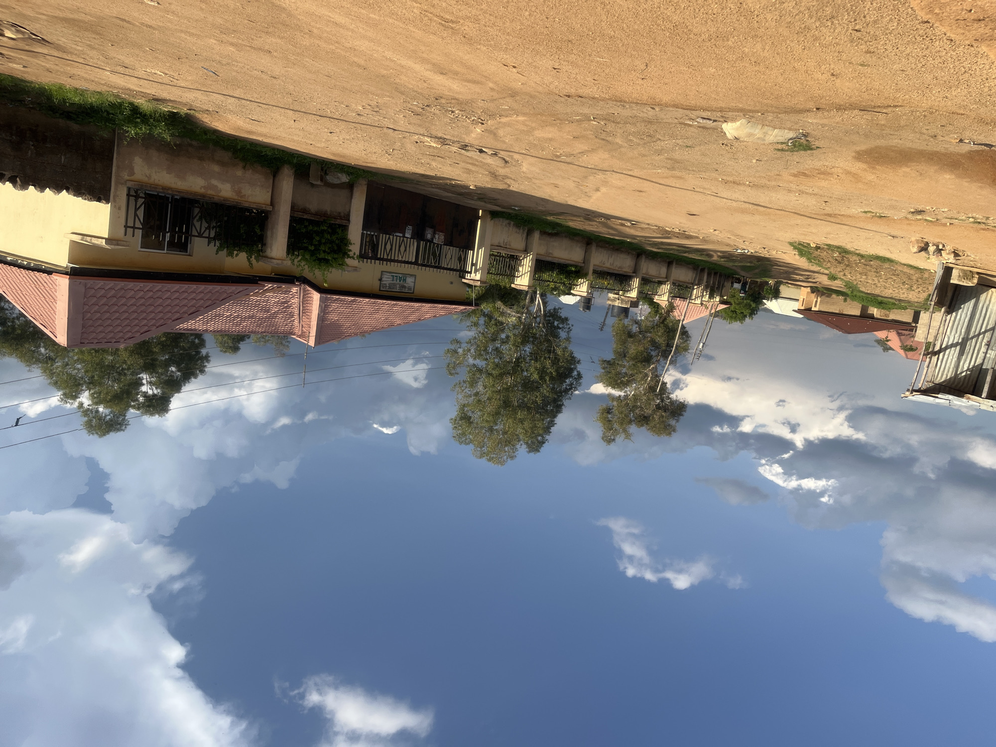 The image depicts an unpaved, reddish-brown dirt road in Barakin Akawo, Nigeria, under a partially cloudy blue sky with both white cumulus and darker grey stratocumulus clouds. The scene is illuminated by natural light, casting subtle shadows.
On the left side, a dilapidated or unfinished structure with corrugated metal siding is partially visible. The dirt road curves upward and to the left, revealing distant residential buildings with reddish-brown roofs. An embankment with sparse green vegetation lines this section of the road.
The center of the image is dominated by the wide dirt road, which shows evidence of vehicle tracks and uneven terrain. In the mid-ground, two tall trees with dense green foliage stand against the sky.
On the right side of the road, a concrete wall topped with a black metal fence encloses a property. A prominent, single-story building painted yellow with a red tiled roof is situated behind this fence. A large, dark brown metal double-gate, likely the entrance to the property, is visible. Above the gate, a white sign with black text reads "THE REDEEMED CHRISTIAN CHURCH OF GOD" at the top, followed by "PRAYER HALL" and "SUNDAY SERVICE" with "8:00 AM" below it, identifying the building as a church hall. Further along the right, another section of the yellow building features a window secured by a black metal grille, and the perimeter wall continues with some green foliage. Utility wires traverse the upper right portion of the sky. In the far distance on the road, two indistinct figures are visible, one appearing to wear a light-colored top, moving away from the prominent yellow building.