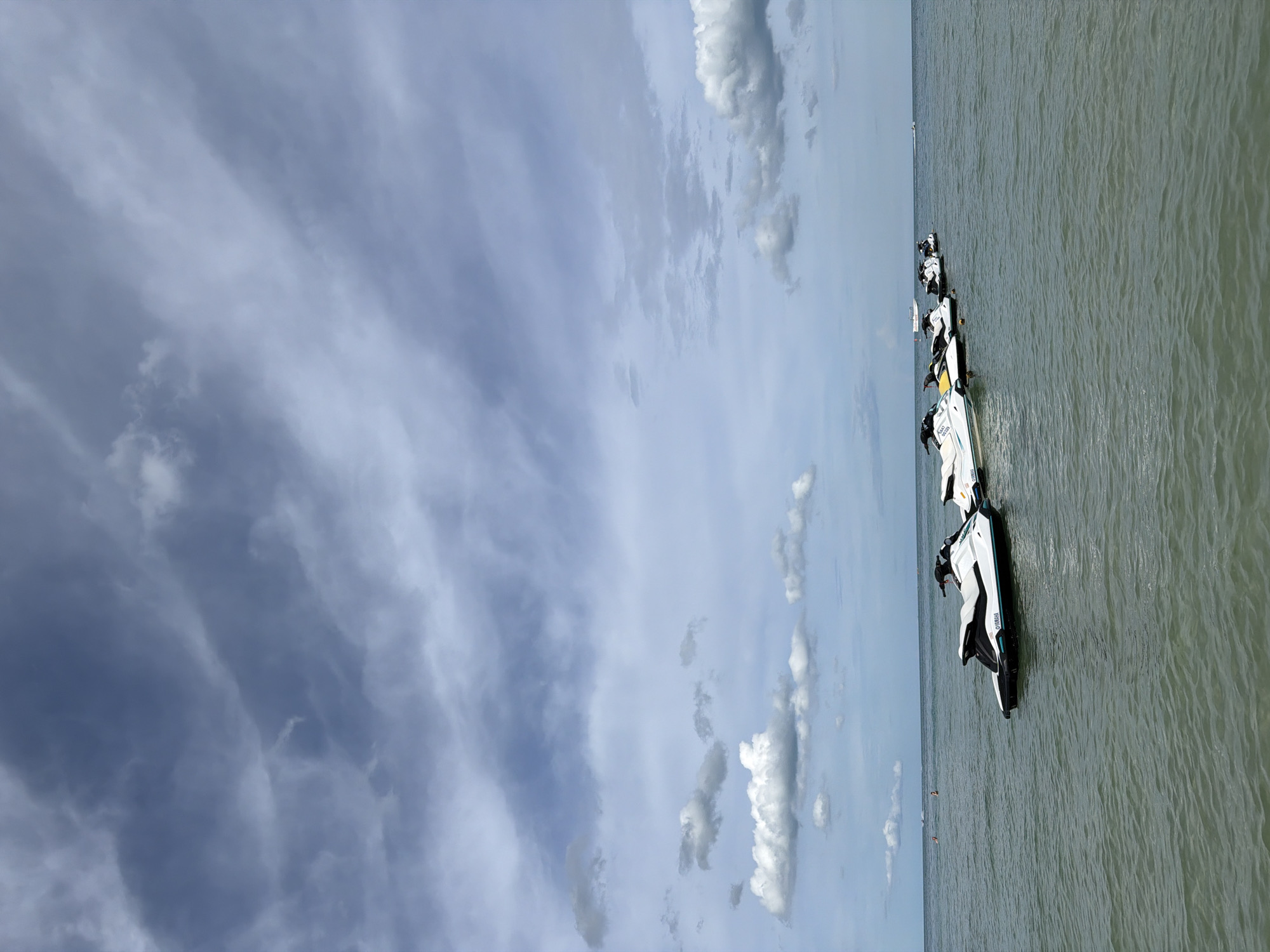 A line of Yamaha jet skis are anchored in the ocean off the coast of Baan Chaweng, Thailand. The sky is overcast with dramatic grey and white clouds, and the water is a murky green. The jet skis are white with black trim and appear to be unoccupied. In the distance, a few small boats are visible on the horizon.