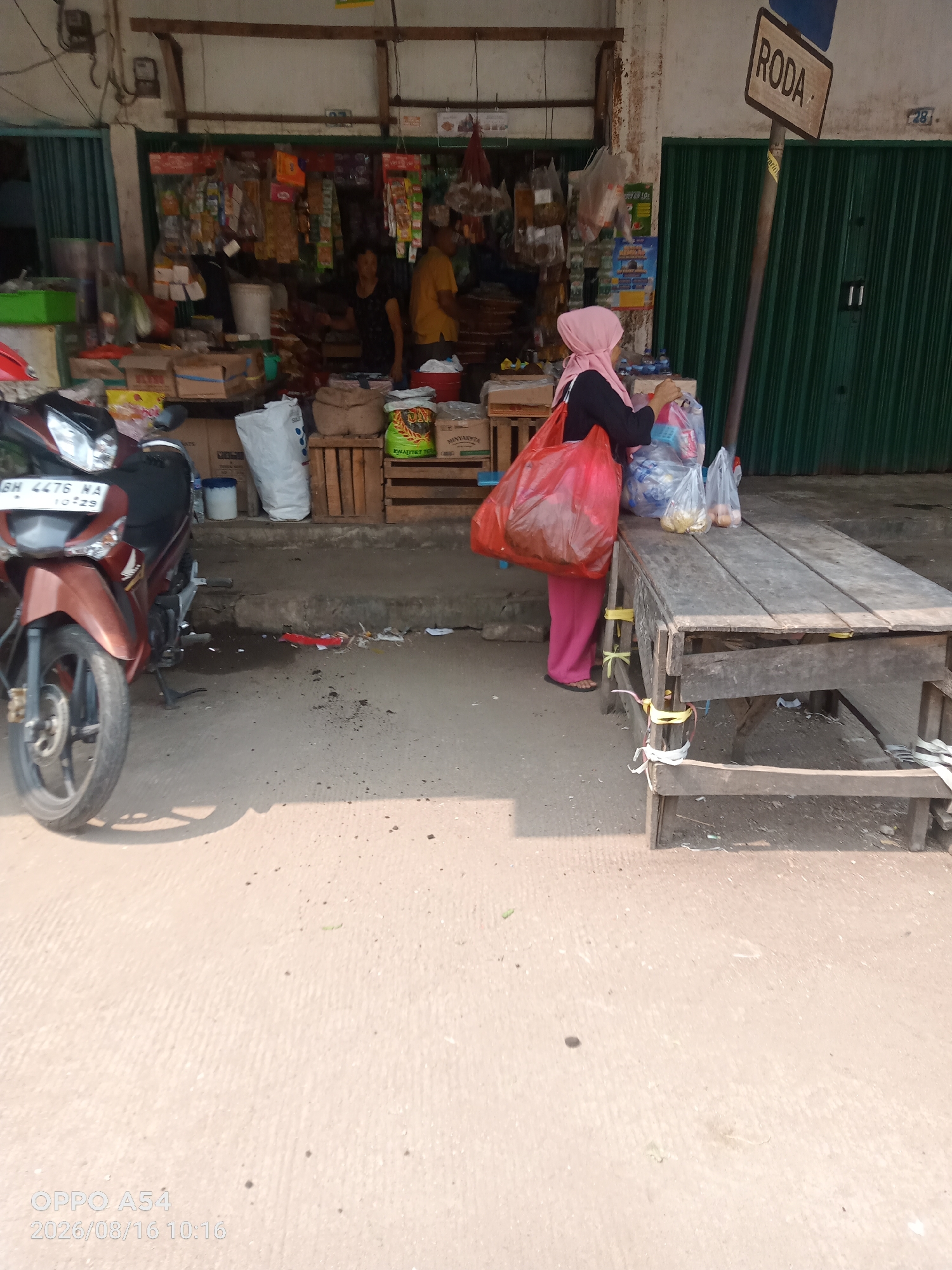 In the city of Jambi, Indonesia, a woman wearing a pink hijab and pink pants is standing next to a wooden table outside a shop. She is holding a large red plastic bag in her left arm and several plastic bags filled with items in her right hand. The shop behind her has green metal roll-up doors and displays various goods on shelves. A motorcycle, brown in color, is parked to the left of the scene. A wooden sign with the word "RODA" is visible on a pole to the right of the woman. The ground is paved with concrete. The timestamp "2026/08/16 10:16" and "OPPO A54" are visible in the bottom left corner of the image.