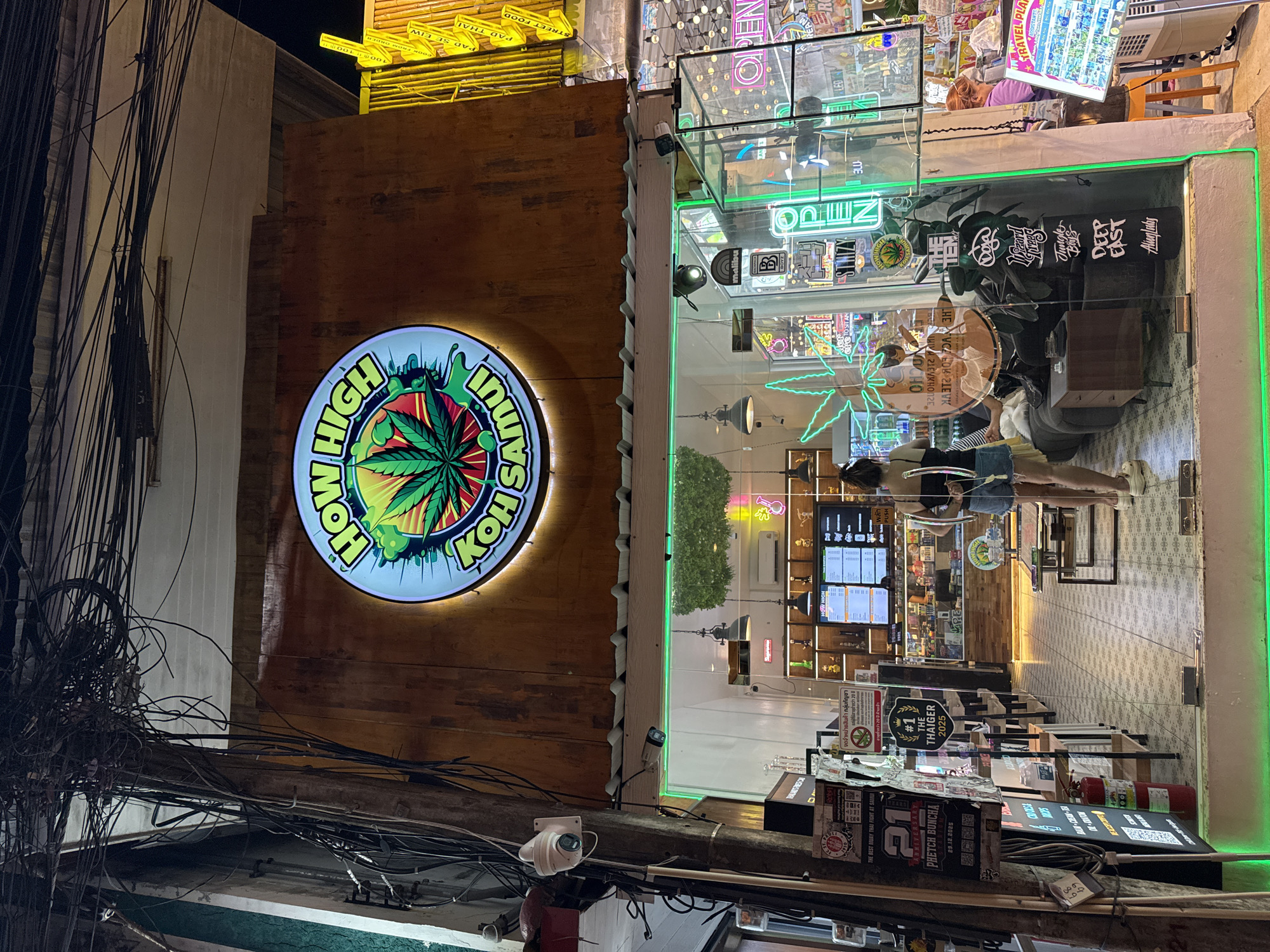 A nighttime exterior shot captures a cannabis dispensary called "How High Koh Samui" in Baan Bo Phut, Thailand. The storefront is illuminated by neon green lighting along the bottom and a large, circular sign above the entrance featuring a cannabis leaf and the dispensary's name. Inside, shelves are stocked with products, and a menu board is visible on a screen. A woman in a black tank top and denim shorts stands near the entrance, facing away from the camera, with her hands on a glass barrier. Another person, possibly an employee, is seated at a counter inside the store. The street outside is dark, with utility poles and tangled wires overhead, typical of urban infrastructure. The overall scene suggests a lively, commercial area at night.