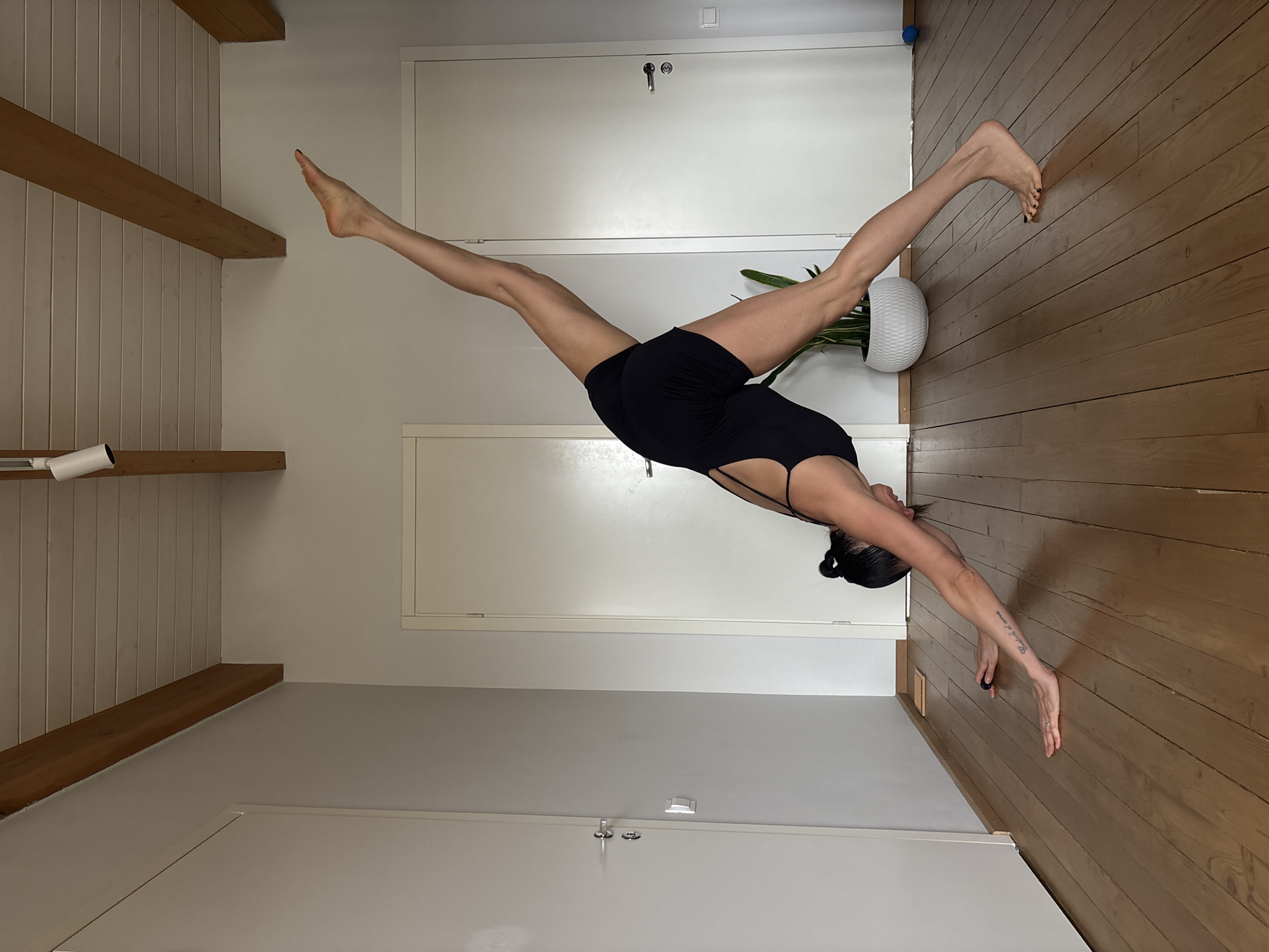 A woman in a black outfit is performing a yoga pose with her legs spread wide, one leg extended towards the ceiling and the other towards a wooden floor. She is bent over with her hands on the floor. The room has wooden floors and white walls with some wooden beams. There is a door in the background. There is a plant pot visible near the woman's leg. The image is taken from a low angle and is slightly rotated. There is no evidence to indicate the location is in Chaikovsky, Russia.