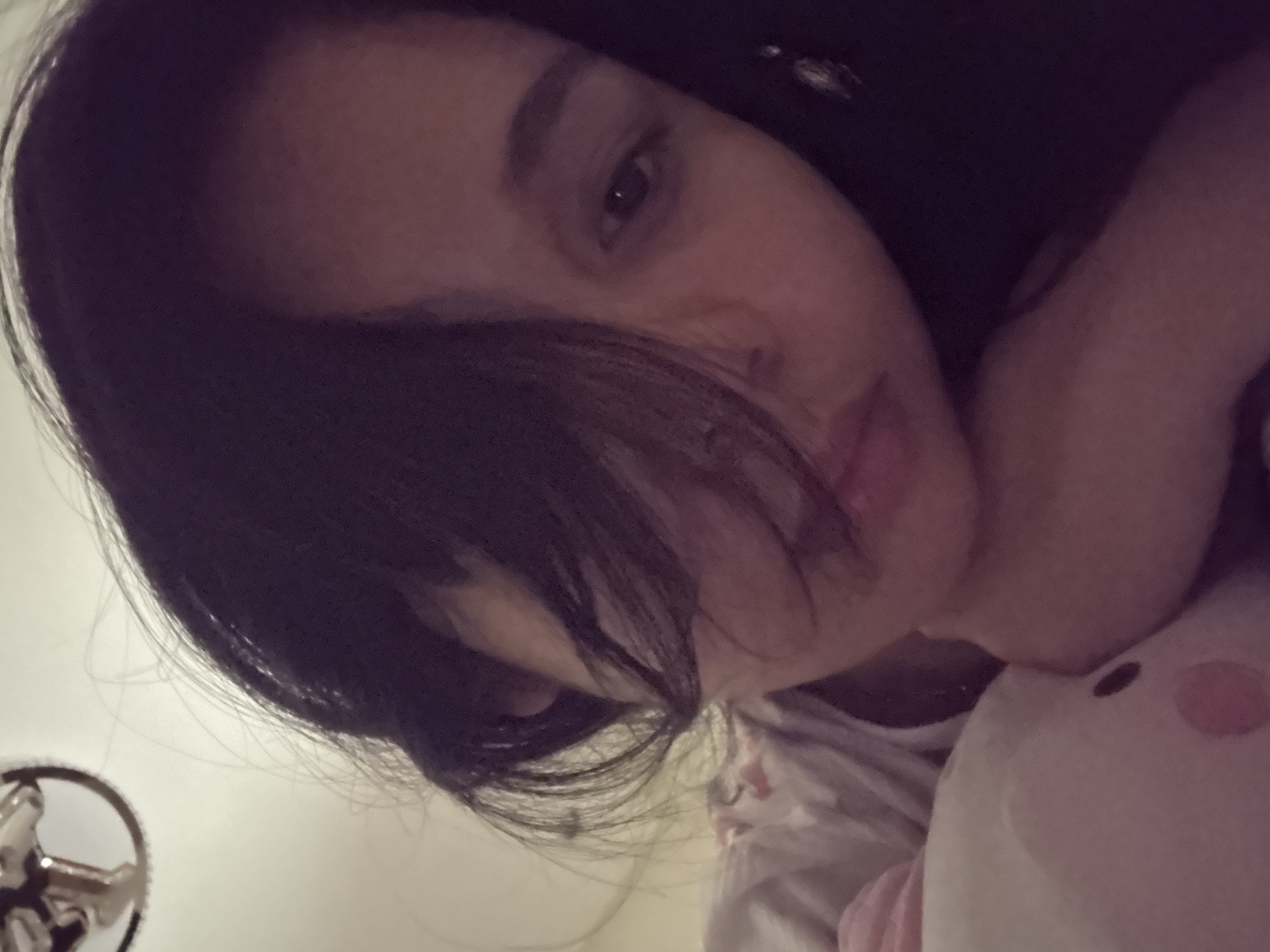 The image displays a close-up, upward-angled portrait of a person, likely female, with long, dark hair partially obscuring their face. The right eye, nose, lips, and chin are visible, while the left side of the face is covered by hair. The person's gaze is directed slightly downward. They appear to be resting or lying down, with one arm or hand positioned near a light-colored, possibly white or pale pink, plush toy that resembles a pig, visible in the lower left quadrant of the frame. The toy has a small dark circular detail and a larger pink circular mark. The person is wearing a light-colored garment with a faint pattern, visible on their shoulder/chest area. The background is a plain, light-toned ceiling or wall, out of focus. A metallic, chrome-colored object, consistent with a ceiling fan blade or light fixture, is partially visible in the upper left corner. The lighting is dim and warm, casting a slightly reddish hue over the scene, suggesting an indoor evening or low-light setting. There are no discernible elements within the image that provide specific geographic information, such as landmarks or identifying cultural cues, to confirm a location in Davao City, Philippines.