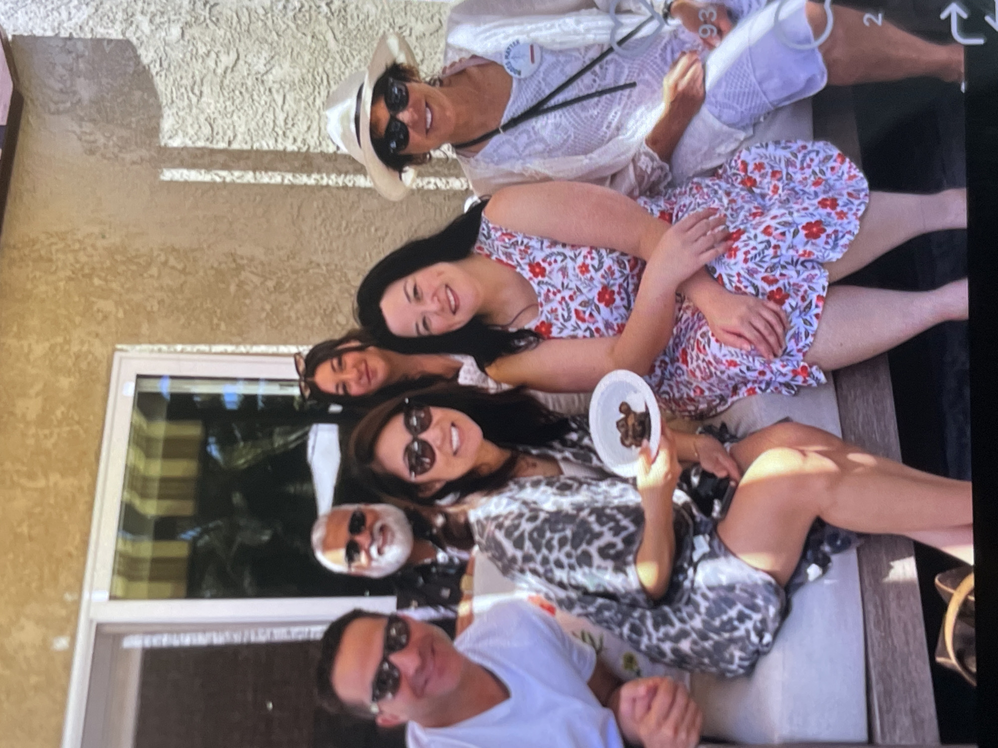 Six individuals are seated on an outdoor sectional sofa. A man in a white t-shirt and sunglasses is seated on the far left, followed by a woman in a patterned kimono, holding a small plate with a piece of chocolate. Behind them, a man with a white beard and sunglasses smiles. Next to the woman with the plate is another woman in a floral dress. To her right, a woman in a white hat and glasses wears a white lace cardigan over a pink top. She has a button pinned to her cardigan. The individuals are posing for a photo in an outdoor setting with a stucco wall and a window with blinds behind them.