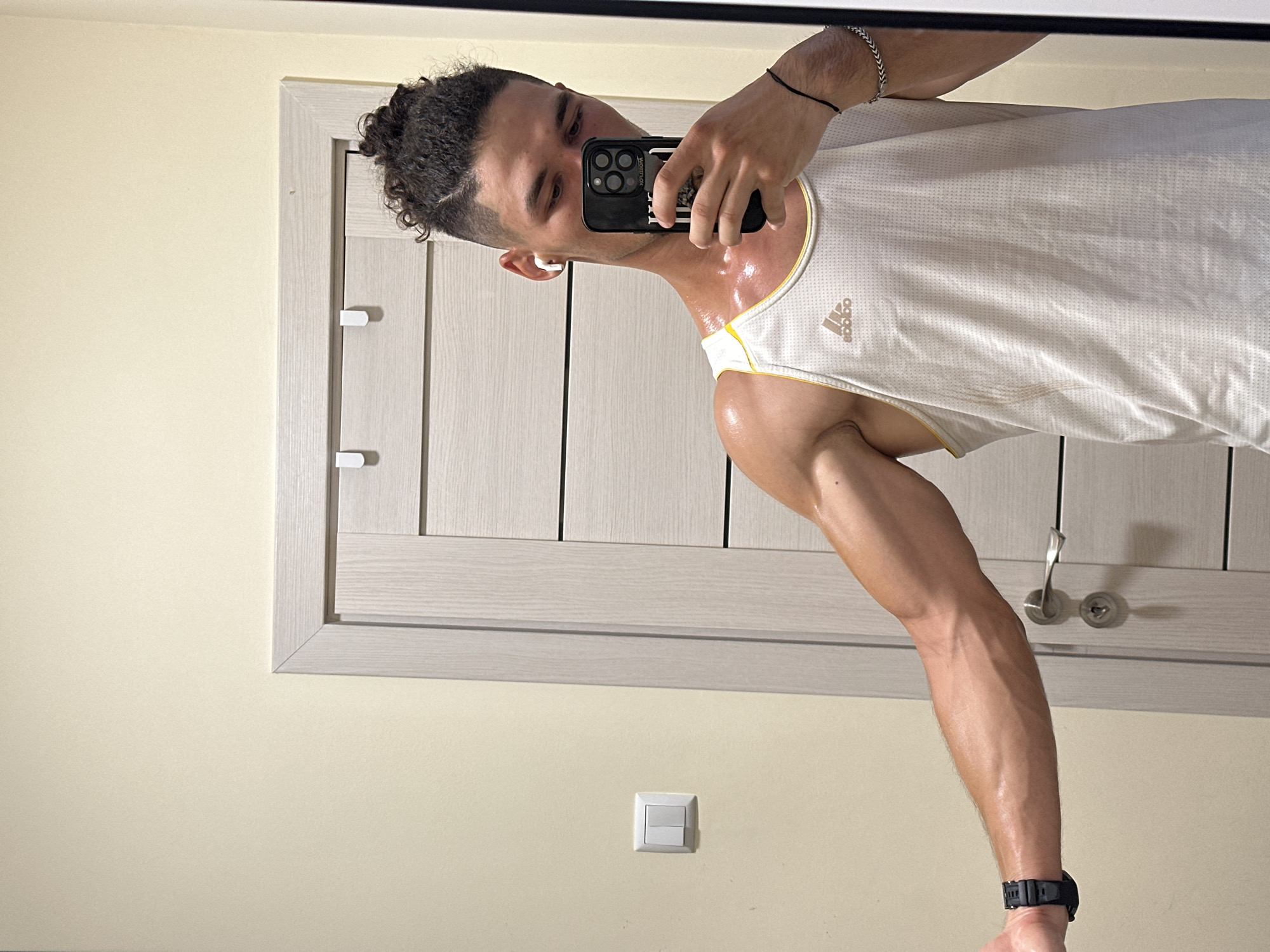 A fair-skinned man with curly brown hair pulled into a bun is taking a mirror selfie in a room. He is wearing a white Adidas tank top, an Apple Watch on his left wrist, and white wireless earbuds. The man is sweating and flexing his left arm, which is highly vascular. In the background, a beige wall is visible, with a light switch on the left side and a light gray door with horizontal panels and a silver handle on the right. The room appears to be a domestic interior, possibly a bedroom or bathroom, in an unknown location.