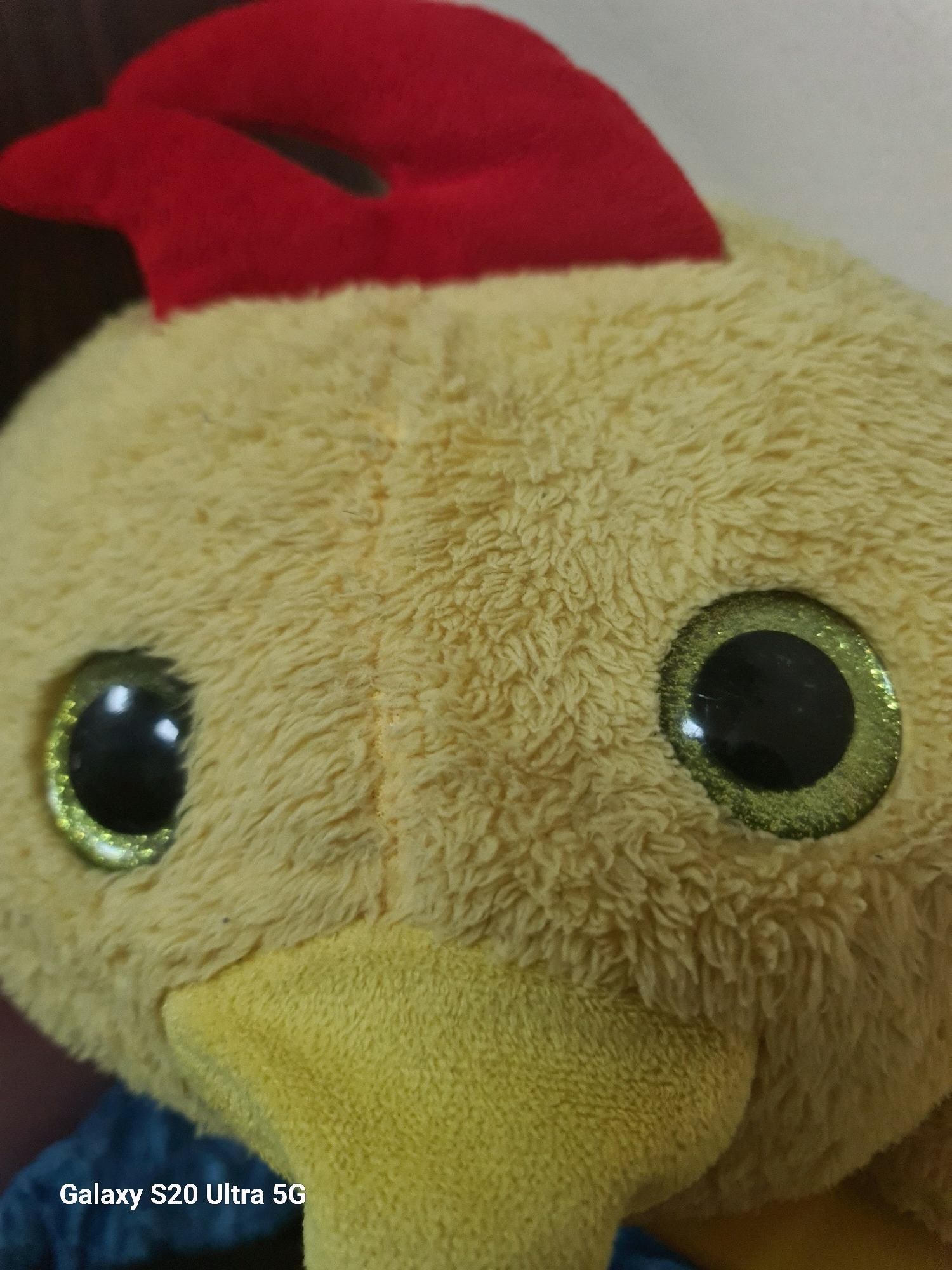 A close-up, head-on, eye-level shot focuses on the face of a yellow plush toy chicken, with a red comb on its head. The toy is positioned in the foreground, occupying the majority of the frame. The background is dark and out of focus. The texture of the plush toy's fabric is visible, with individual fibers appearing soft and fuzzy. The chicken's eyes are large, black, and round, with a sparkly green outer ring. Its beak is a lighter yellow and appears to be made of a different material, possibly felt. The image is captured indoors, with artificial lighting. There is no discernible activity or interaction taking place. No people are visible in the image. The photograph contains a watermark in the bottom left corner that reads "Galaxy S20 Ultra 5G". The location in the city of Sohar Province, Oman, is not discernible from the image itself.