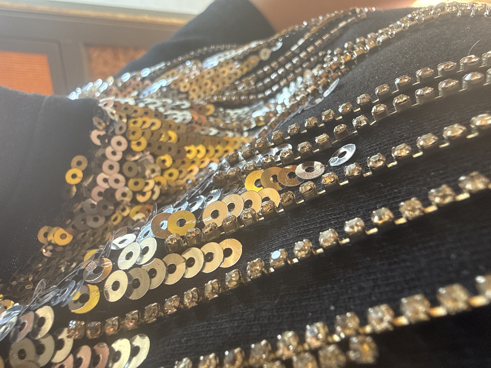 The image shows a close-up view of a piece of dark fabric embellished with rows of sequins and rhinestones. The sequins are primarily gold in color, while the rhinestones appear to be set in a metallic base. The arrangement of the embellishments creates a patterned or decorative effect on the fabric.