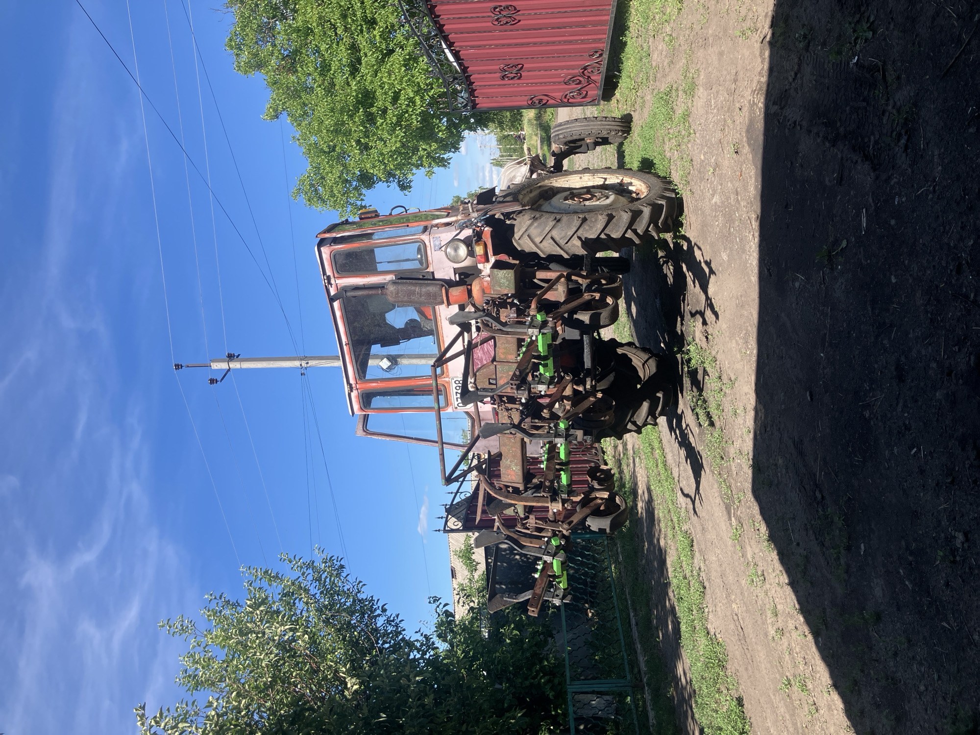 An aged, red tractor with a multi-row plow attachment is parked on a dirt path in Vovkivka, Ukraine. The tractor has a metal cab with visible windows and a driver inside, partially obscured. The plow attachment features several metal blades with green protective covers. The tractor is positioned in front of a tall, red metal fence with decorative scrollwork. To the left of the tractor, a green metal fence runs alongside a lush green tree. In the background, a residential building with a light-colored roof and a utility pole with electrical wires are visible. The sky is clear and blue with scattered white clouds. The ground is a mixture of dirt and sparse green grass. Shadows from a nearby structure and the tractor are cast on the ground.
