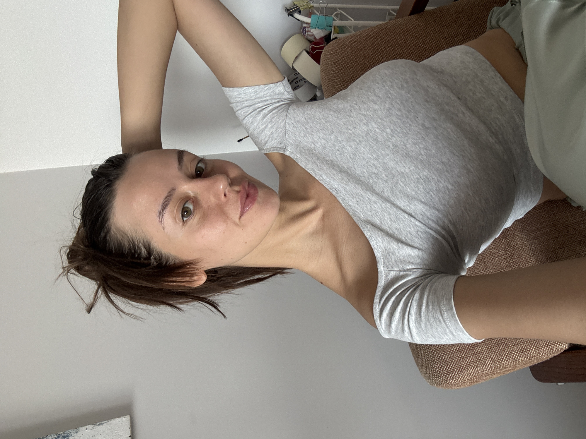 A young adult female is positioned centrally, taking a selfie. She is seated in a light brown upholstered chair, with her right arm (viewer's left) bent and raised behind her head, and her left arm (viewer's right) resting near her side. She looks directly at the camera with a subtle smirk or slight smile. Her dark brown hair is pulled back in a messy ponytail or bun, with loose strands around her face and forehead. She wears a light gray, short-sleeved, off-the-shoulder cropped top, exposing her midriff. Light green or teal colored bottoms, possibly sweatpants or lounge pants, are visible at the bottom right. Her skin tone is fair, with visible freckles or small moles on her face and neck. Her eyes are brown, and her lips appear glossy.
The background to her left consists of a plain, light gray wall. A fragment of a white abstract artwork with blue markings is partially visible in the upper left corner. To her right, a white wall forms a corner. Behind her and to her right, a white wire or plastic caddy is visible, containing several rolls of adhesive tape (appearing to be masking and packing tape), small containers, and other miscellaneous items. The scene appears to be an indoor, casual home setting.