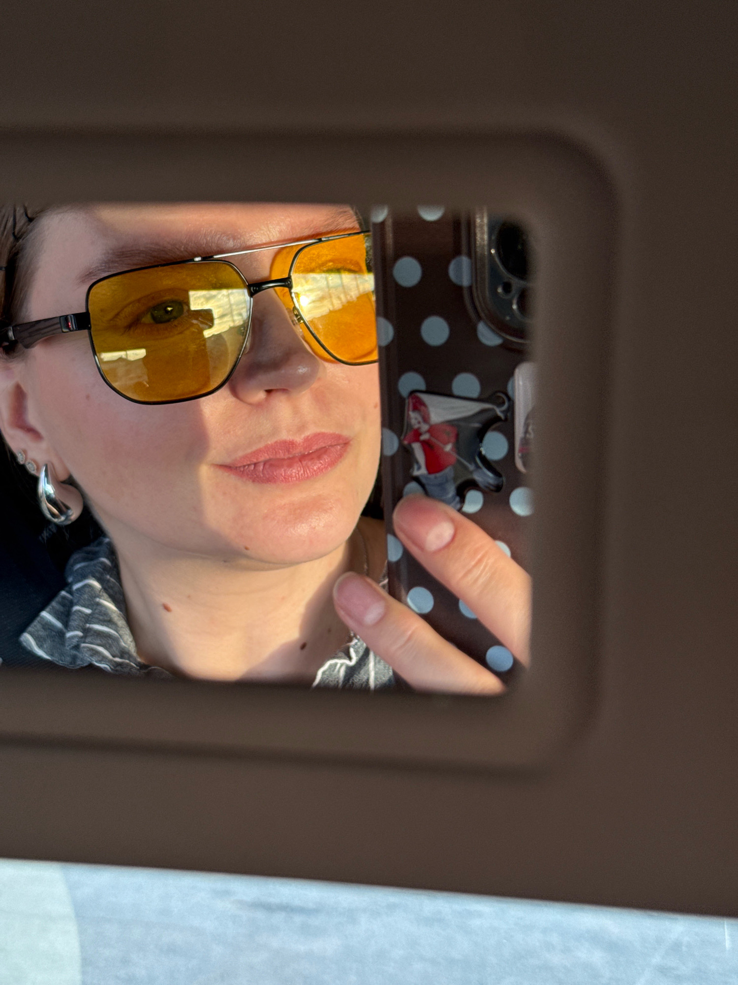 The image shows a selfie of a person wearing yellow-tinted aviator sunglasses and a striped shirt, reflected in a car's visor mirror. The person's left eye is visible through the sunglasses, reflecting sunlight and possibly buildings. A cell phone with a polka dot case is held by a hand, also reflected in the mirror, obscuring part of the scene. The background appears to be an interior of a car, with the visor's shadow casting across the reflection. The location is specified as Aksay, Kazakhstan, but there are no specific visual cues in the image to confirm this detail.
