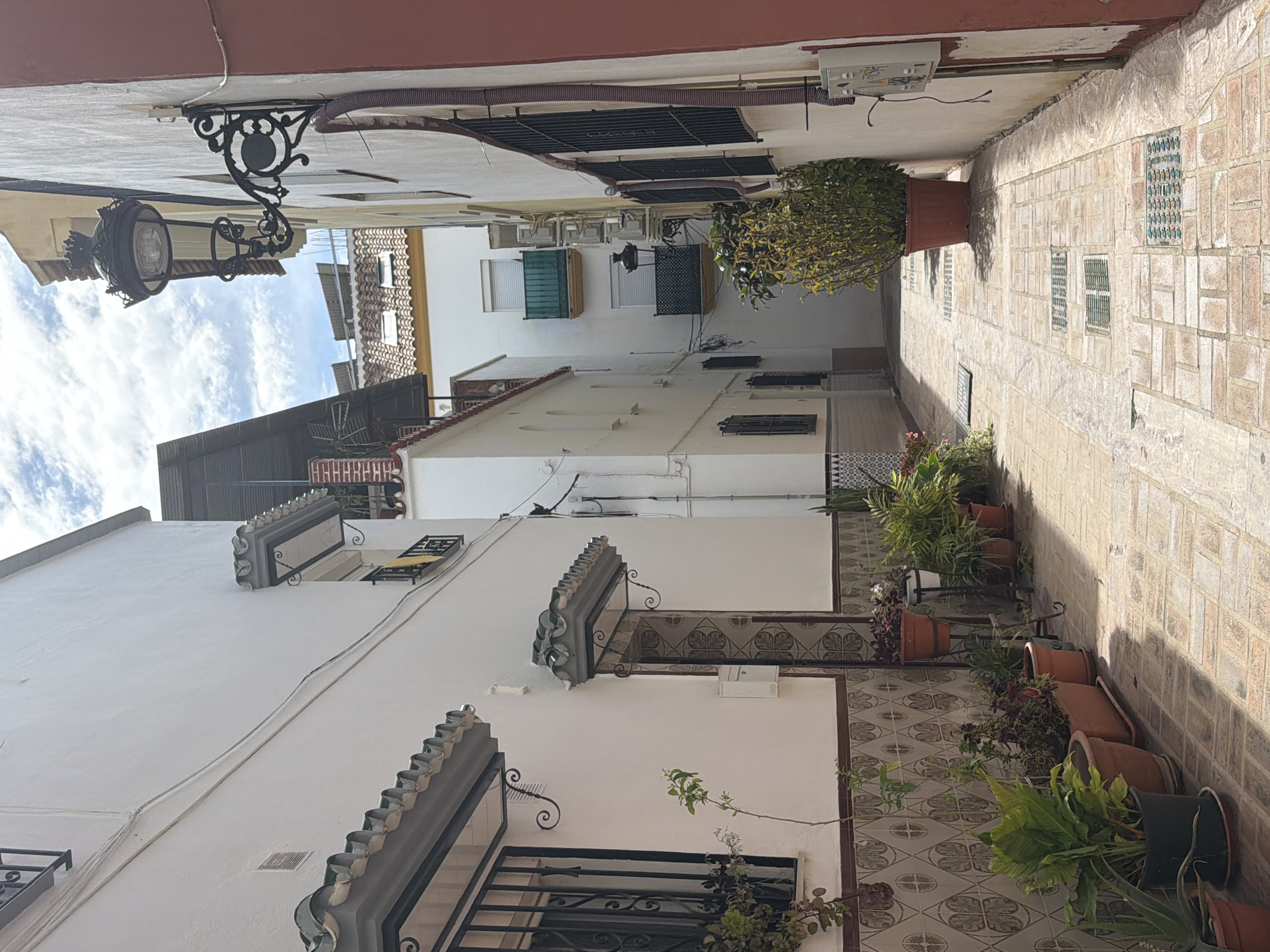 The image captures a narrow alley in Arroyo De La Miel-Benalmadena Costa, Spain. The alley is flanked by buildings, mostly white with some terracotta accents on the roofs and upper walls. Decorative ironwork is visible on some windows and balconies. Potted plants line the base of the walls along the alley floor, adding greenery. The paving stones are light-colored and laid in a pattern. An ornate street lamp is attached to the building on the left side of the frame. The sky is partially visible between the buildings, showing scattered clouds.