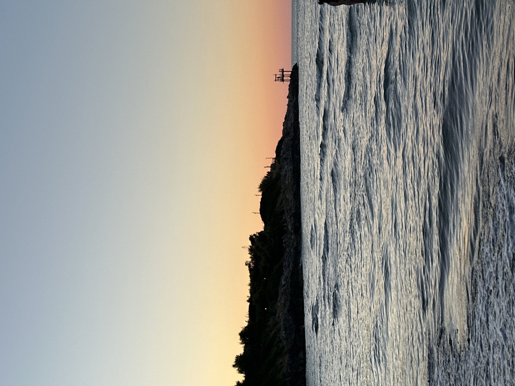 A serene sunset scene unfolds over the Adriatic Sea in Peschici, Italy. The sky transitions from a pale blue at the top to a warm gradient of yellow and orange near the horizon. In the foreground, gentle waves ripple across the water, with some white foam visible closer to the shore.
On the left side of the image, a rugged, tree-covered cliff juts out into the sea. Silhouetted against the colorful sky, the foliage creates a dark, textured line. Perched on a rocky outcrop on this cliff is a distinctive wooden structure, likely a "trabucco," a traditional fishing platform. It features a complex network of beams and a walkway.
On the right side of the image, a person's torso and head are visible from behind, standing in the water. They appear to be facing the sea, observing the sunset. The overall impression is one of tranquility and natural beauty, characteristic of a coastal sunset in Peschici.