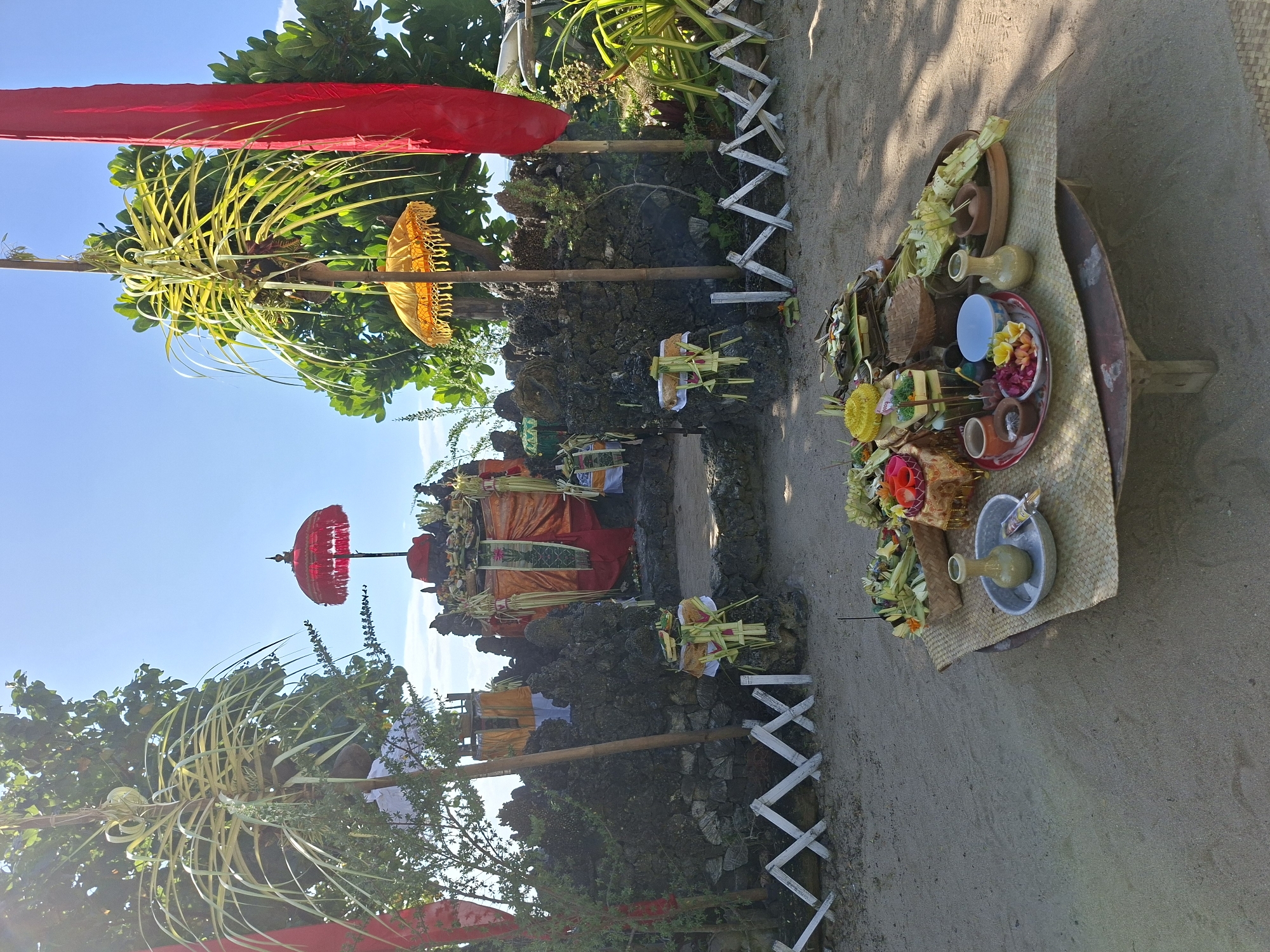 The image shows a Balinese Hindu ritual or ceremony setting. The location appears to be outdoors, possibly on a beach or coastal area, given the presence of sand.
Elements of the scene include:
* **Religious Structures:** What appears to be a small altar-like structure, adorned with orange cloth, sits upon a rocky base.
* **Offerings:** Trays with offerings, potentially flowers, fruits, and other ritualistic objects, are presented on a table covered with a woven mat.
* **Parasols:** Two parasols, one red and one yellow, are used.
* **Penjor**: decorated bamboo poles are erected on either side of the altar.
* **Environment:** The sky is visible, and vegetation is present around the scene.
* **Fencing**: A white, X-shaped fencing is on the ground.
