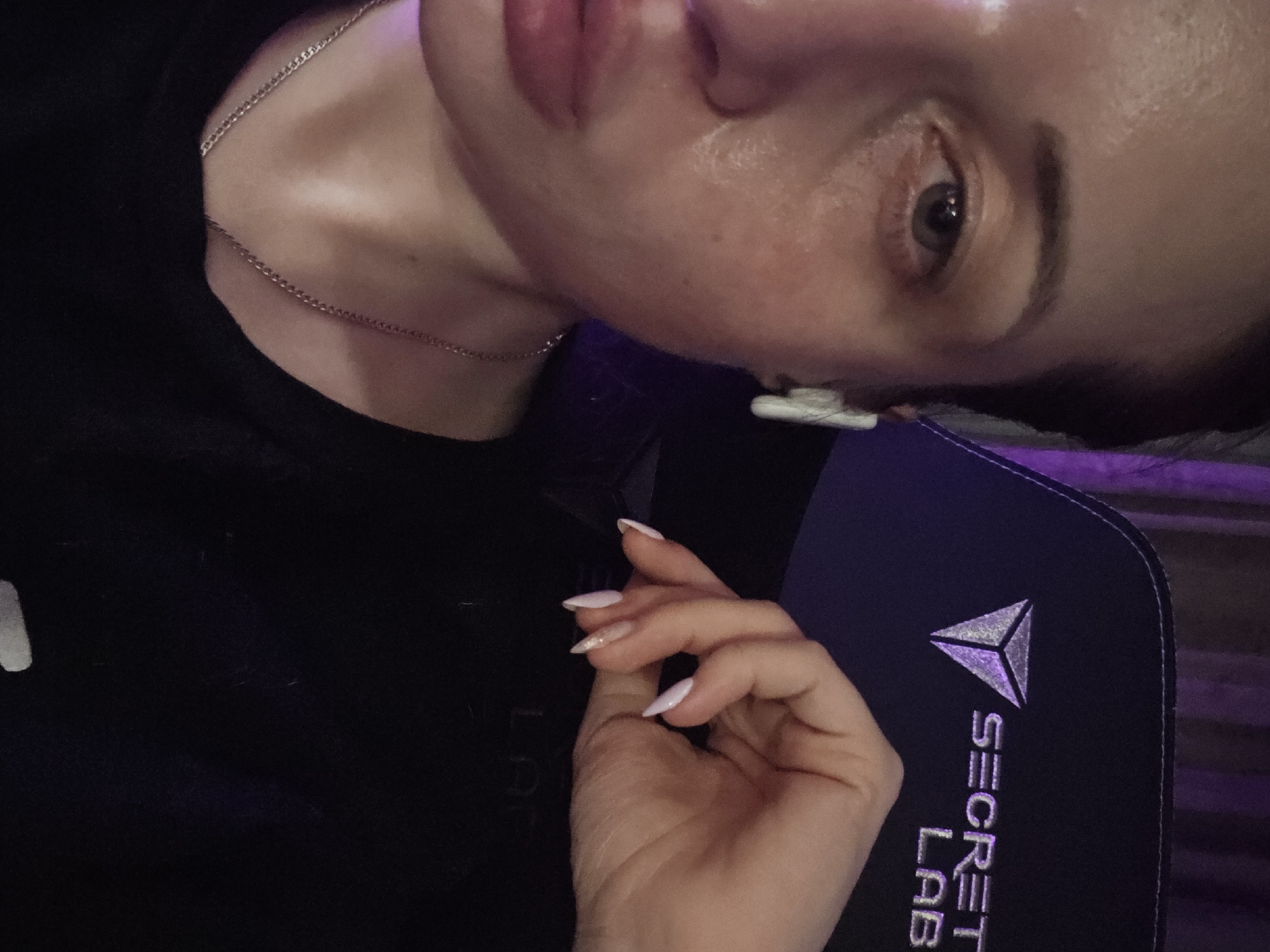 A selfie-style photograph focuses on a person's face and hand. The individual has fair skin, a silver chain necklace, and one visible earbud. Their expression is neutral, with a focus on their eye. Their hand, featuring long, light-colored nails, is positioned near a black Secret Lab gaming chair. The background is dimly lit, suggesting an indoor environment. There is no information in the image that indicates the location of Burgas, Bulgaria.