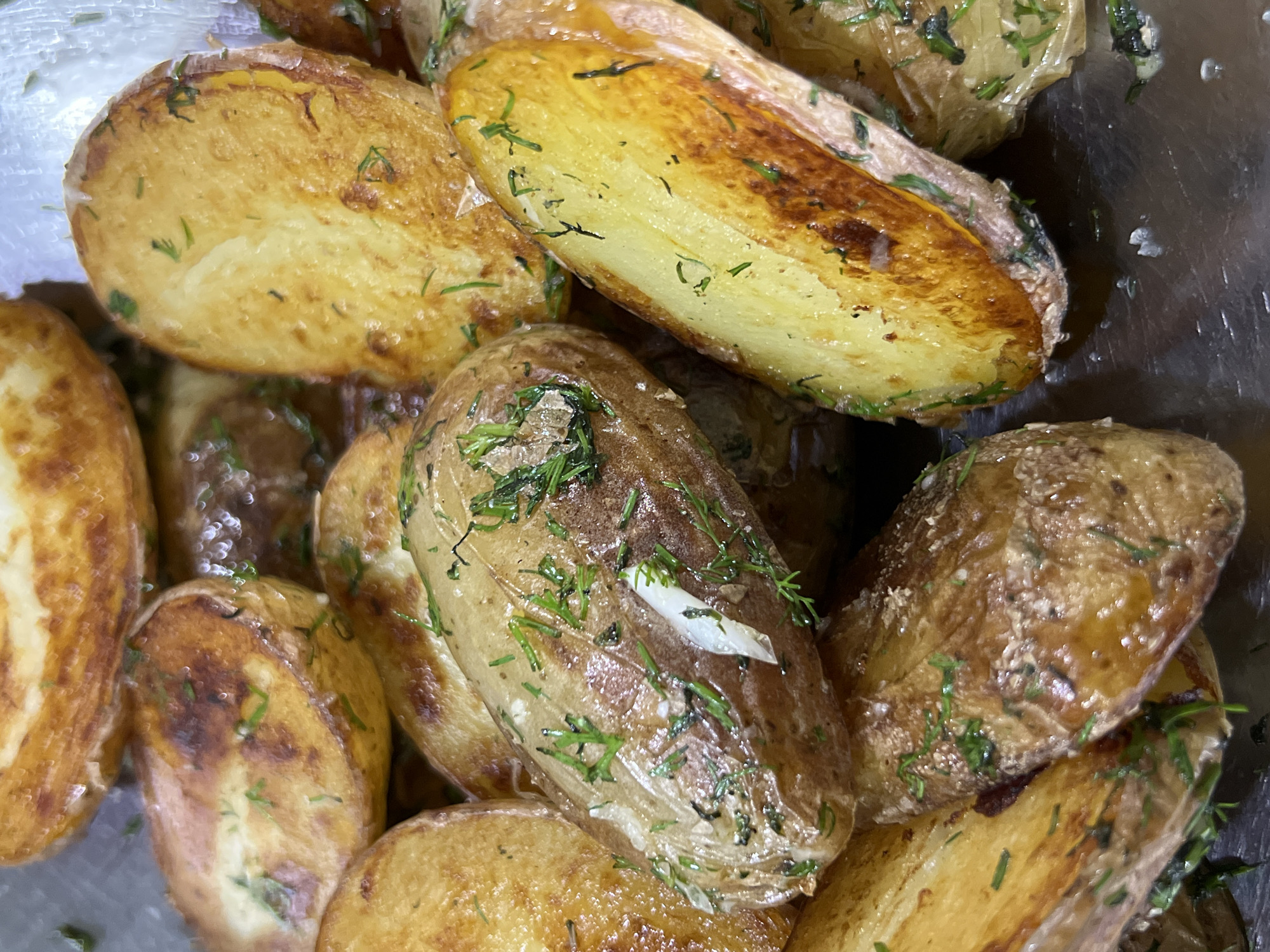 The image shows a close-up, overhead view of a pile of roasted potato wedges seasoned with dill. The potatoes are golden brown and some have darker, crispy edges. They appear to be served in a metallic dish. There is no information in the image to suggest a location in Krasnodar, Russia. The image is a still photograph with no discernible people, actions, or interactions. The environment is limited to the potatoes and the dish they are in.